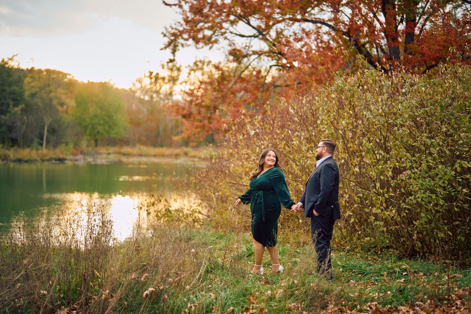 Two people stand by a serene lake during autumn with orange foliage and golden sunlight filtering through the trees.