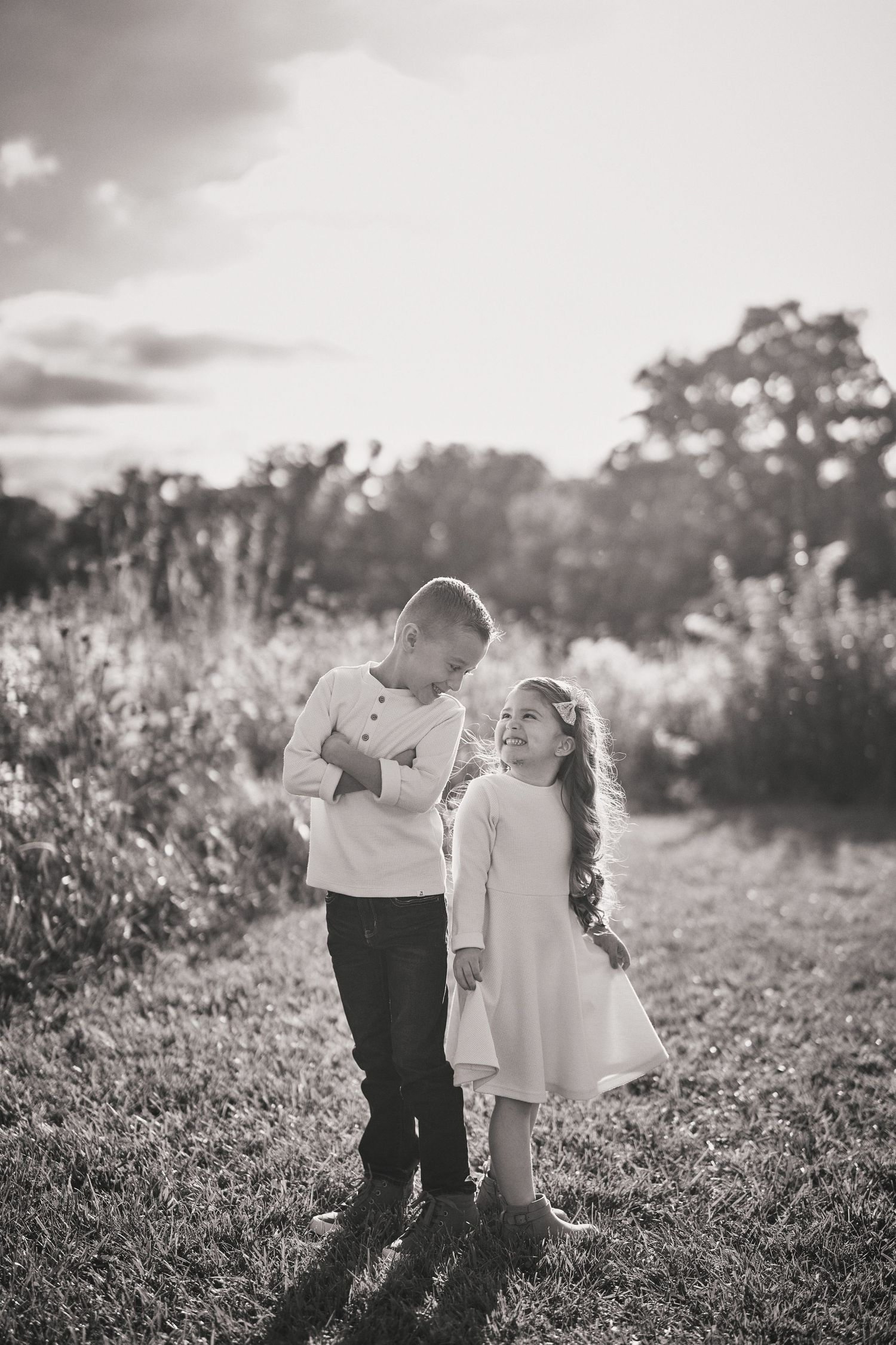 Black and white photo of two children in light colored clothing standing together on a sunlit path in a field at sunset.