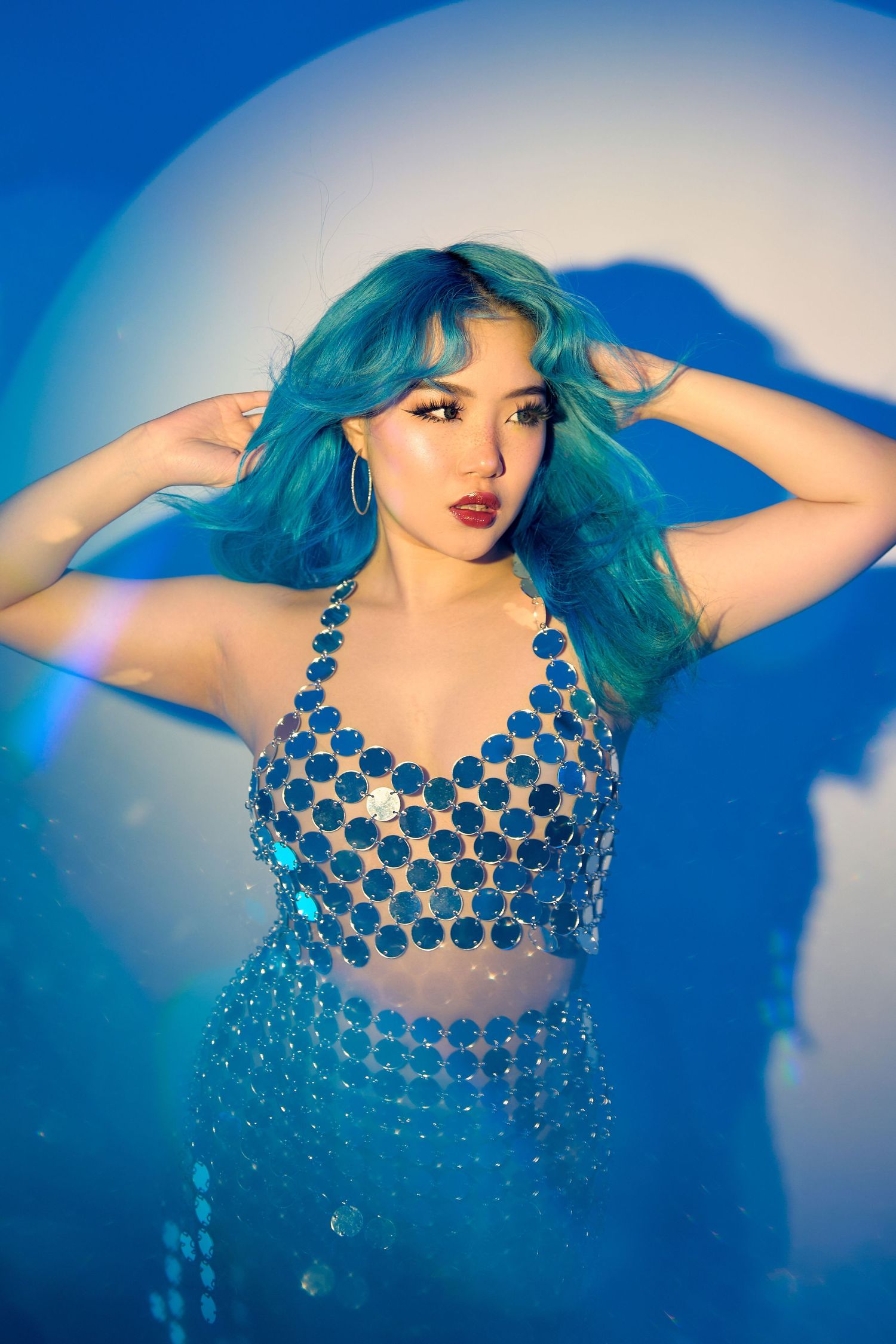 Woman with vibrant blue hair wearing sparkly top posing against blue background.