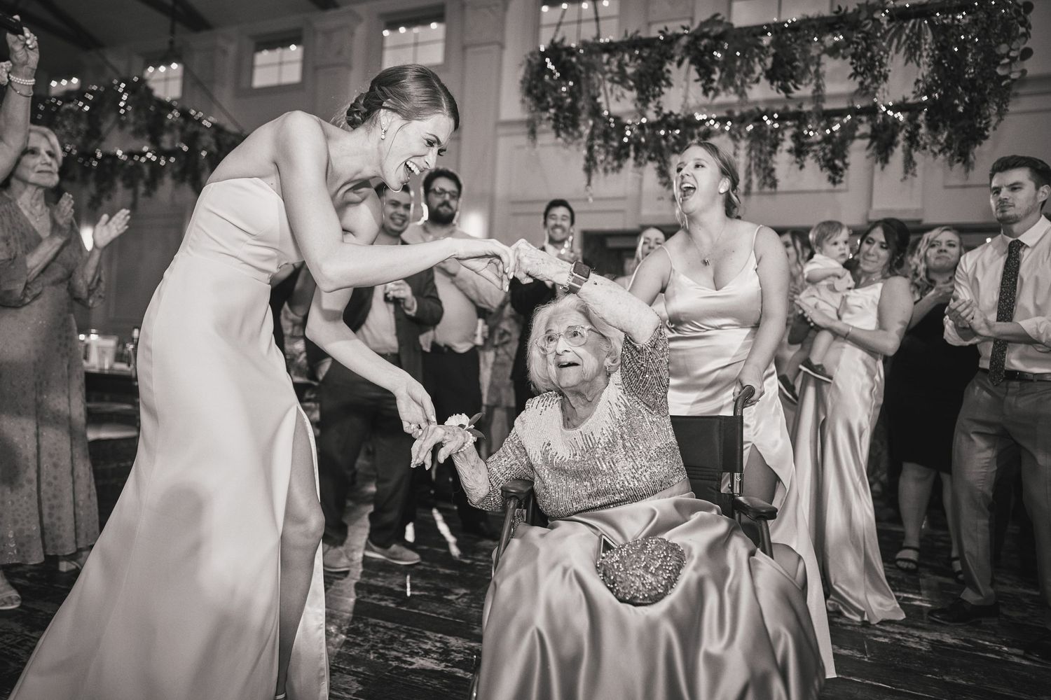 Black and white photo of guests celebrating at a wedding reception as several attendees dance and gather on the dancefloor.