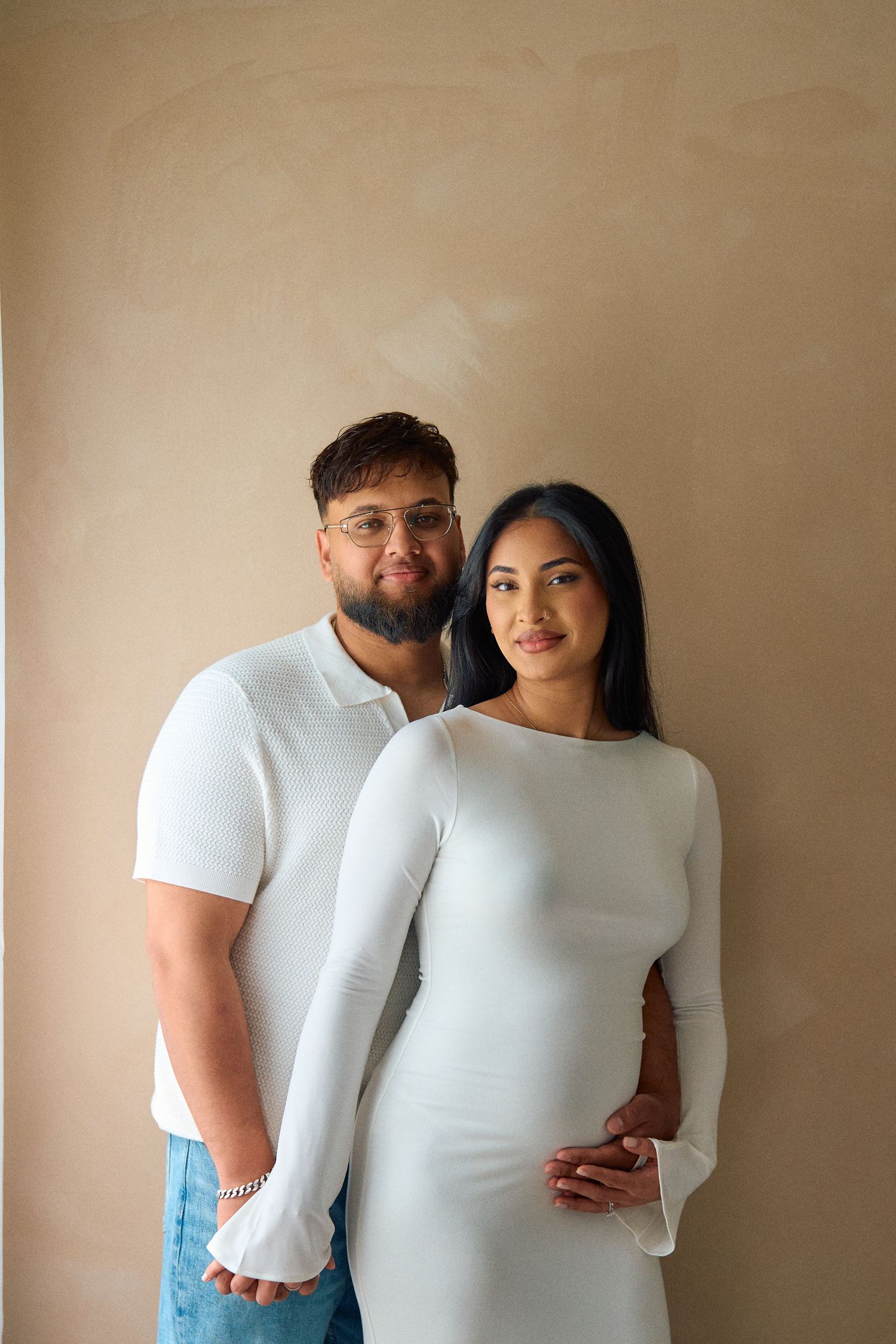 A couple in matching white outfits poses together against a beige wall, with the woman wearing a fitted long sleeve dress.