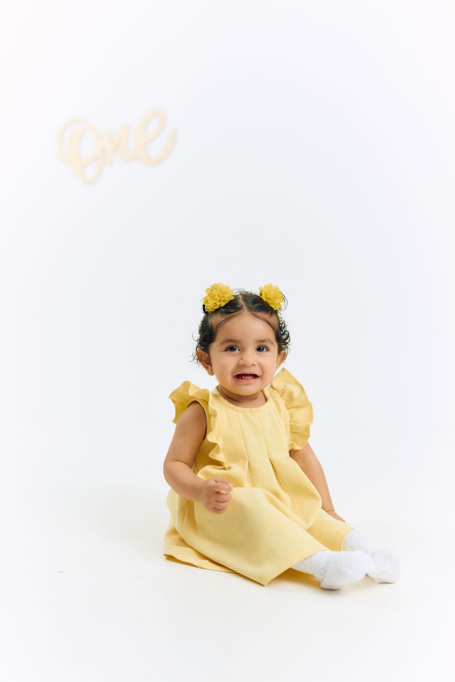 Baby in yellow dress with floral crown sits on white backdrop in three different poses for milestone photo session.
