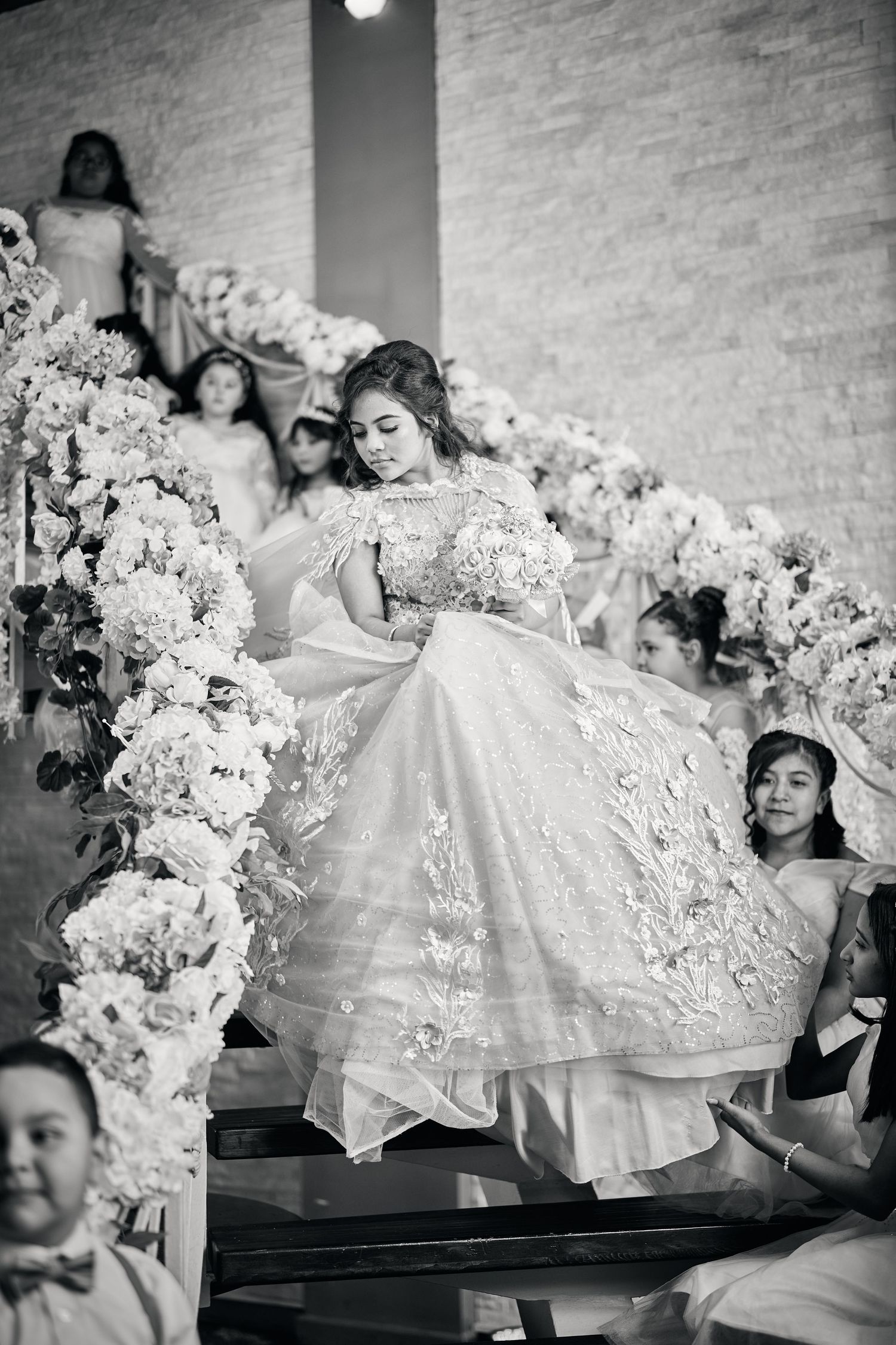 A black and white photo of a bride in an elegant lace wedding gown posing near floral decorations on stairs.