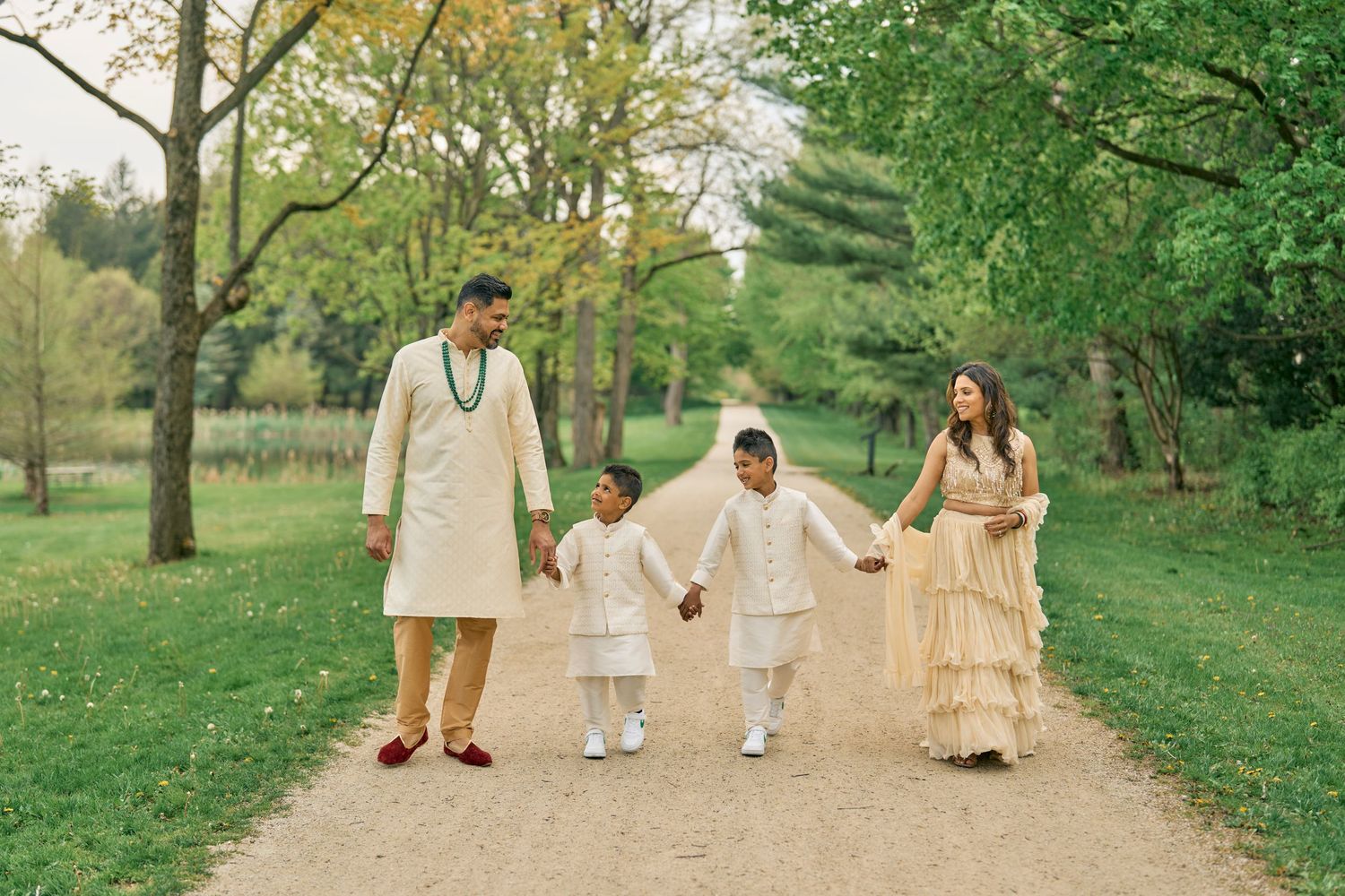 Family in coordinated light-colored outfits walking hand in hand on a tree-lined path in a park during spring.
