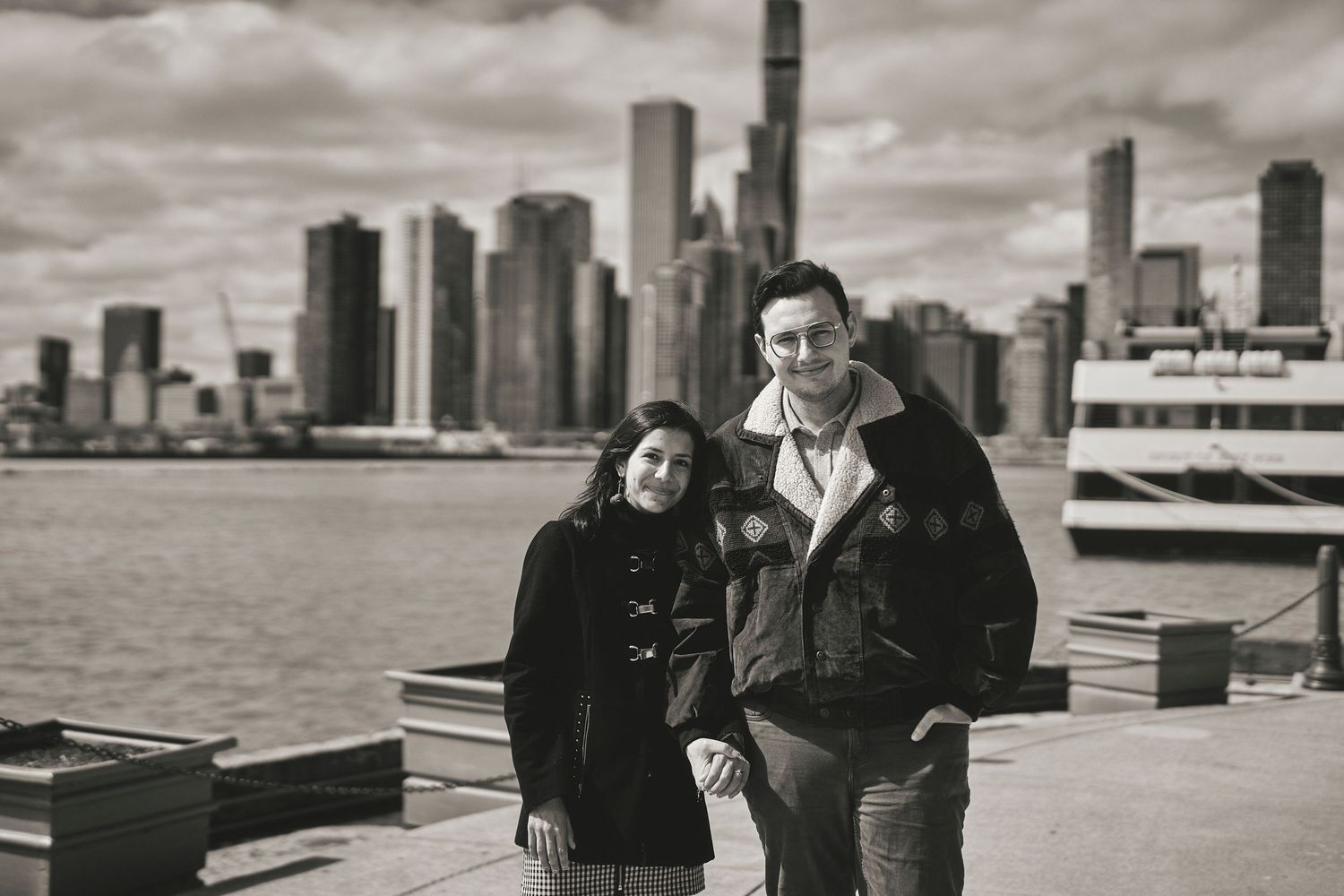 A black and white photo of a couple standing on a pier with the Chicago skyline and a ferry boat in the background.