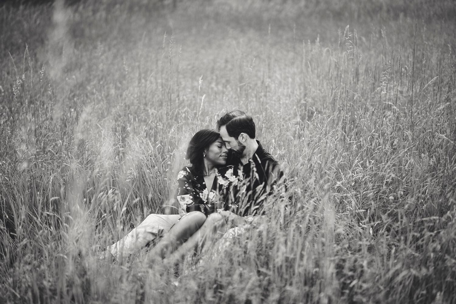 A romantic black and white scene of a couple sitting intimately together in a sun-lit meadow of tall grass.
