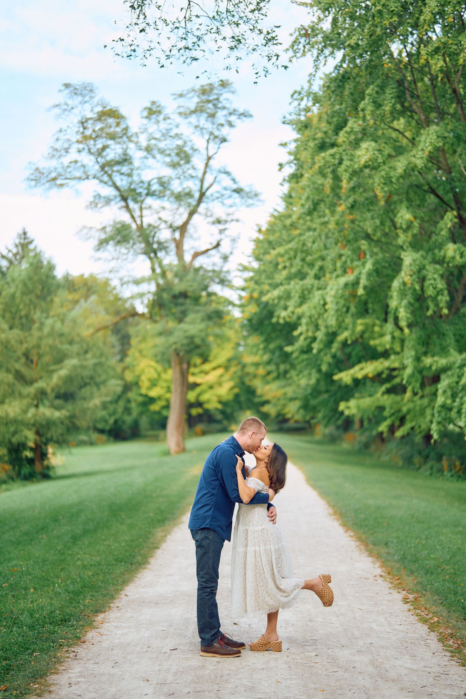 A romantic outdoor moment captured on a tree-lined path with lush greenery in the background on a sunny day.