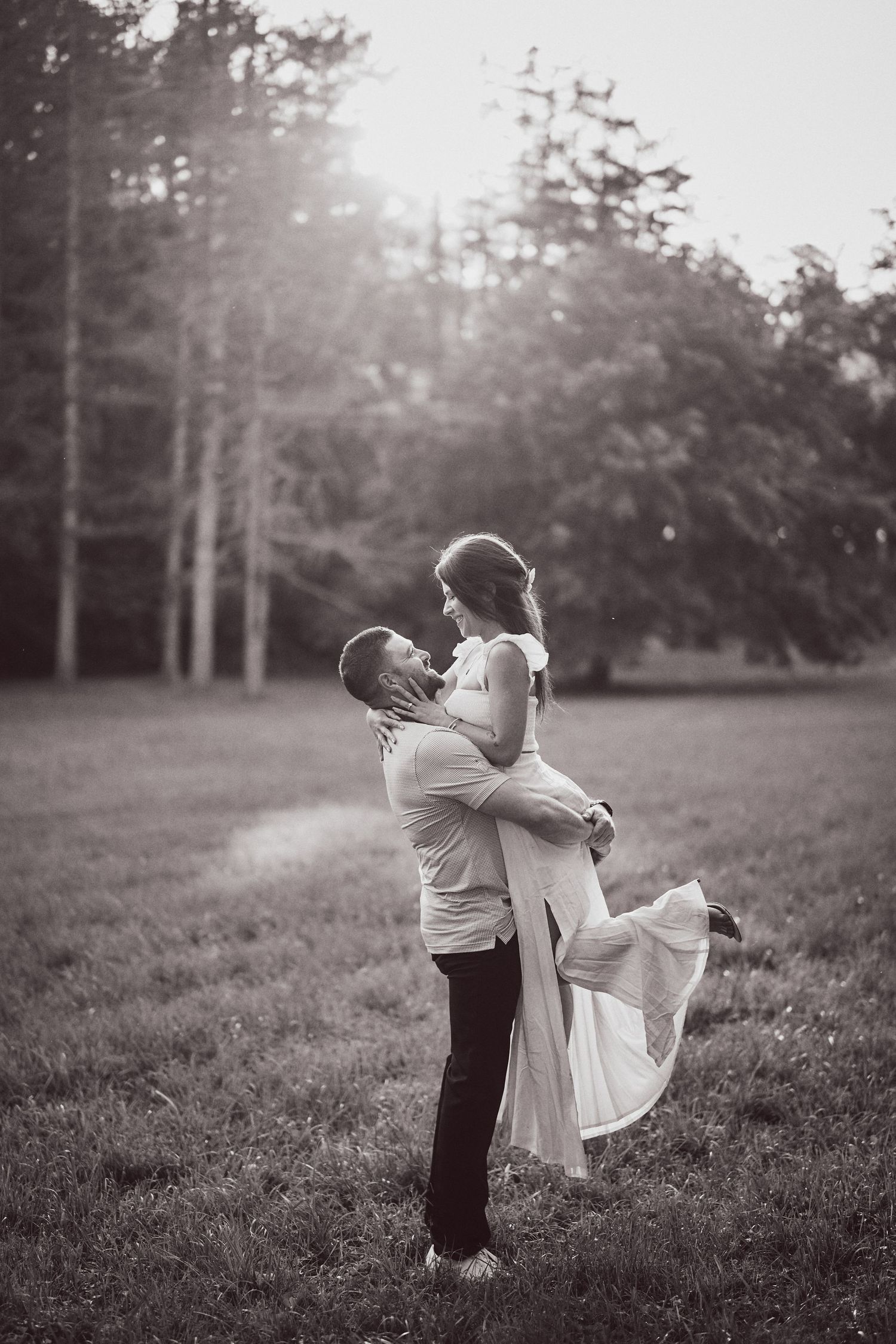 A romantic black and white photo shows a couple embracing in a sunlit meadow with forest trees in the background.