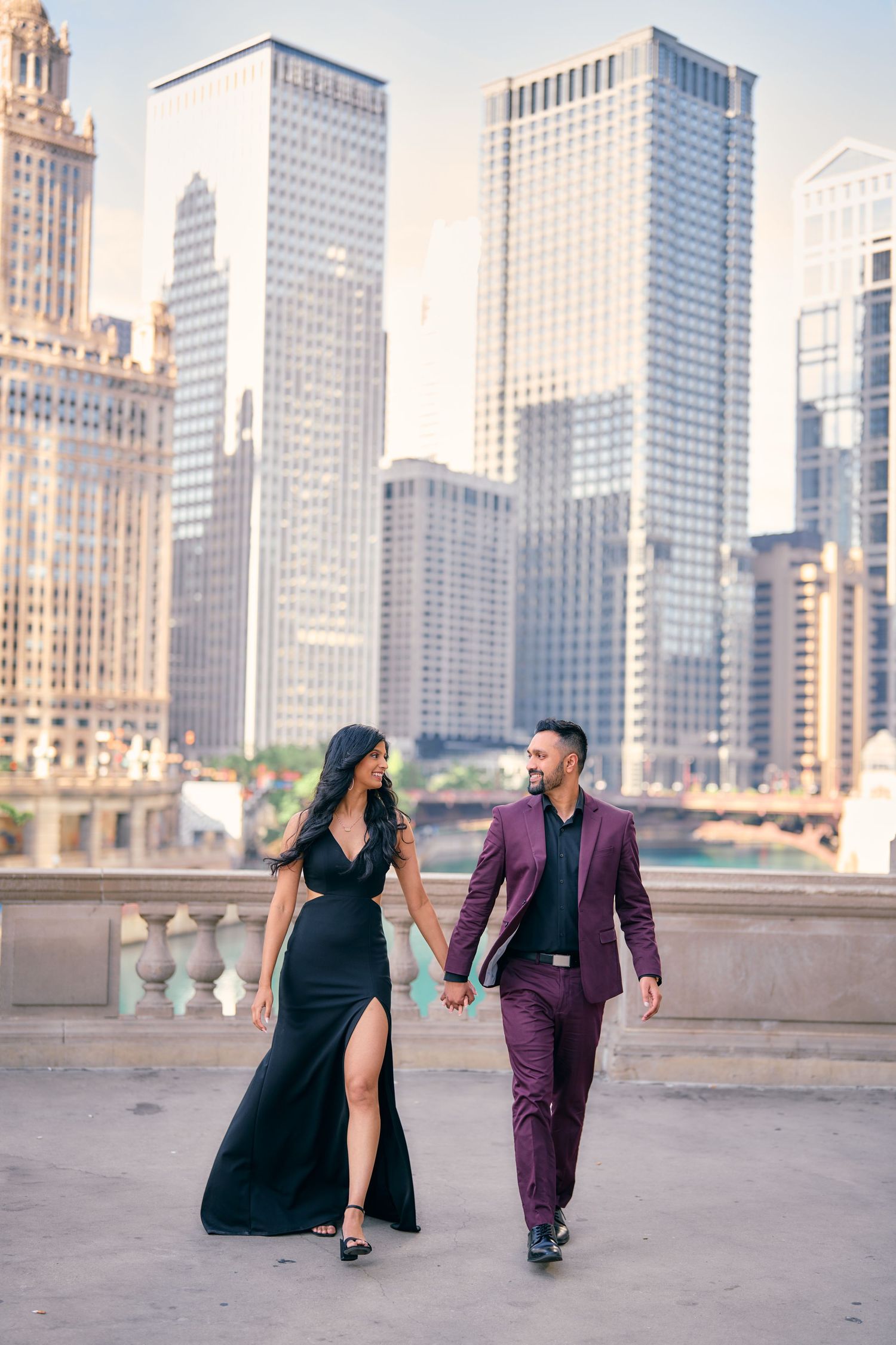 Elegant couple in formal attire walking along Chicago Riverwalk with downtown skyscrapers in background during sunset.