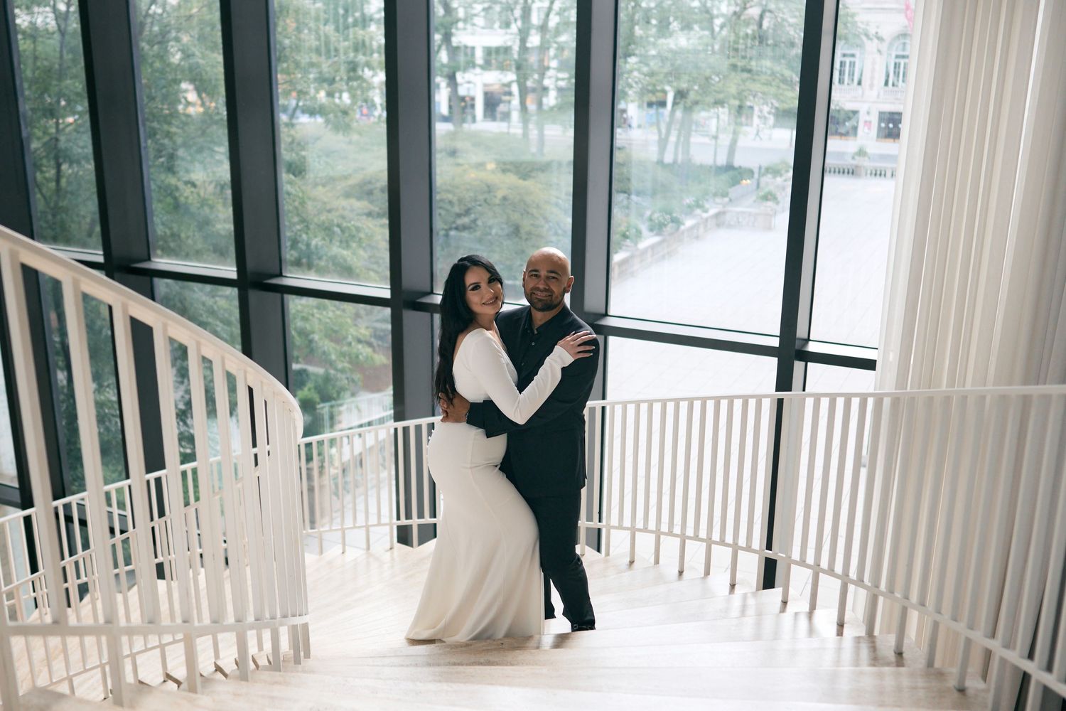 A couple dances together on an elegant white spiral staircase with floor-to-ceiling windows overlooking trees in the background.