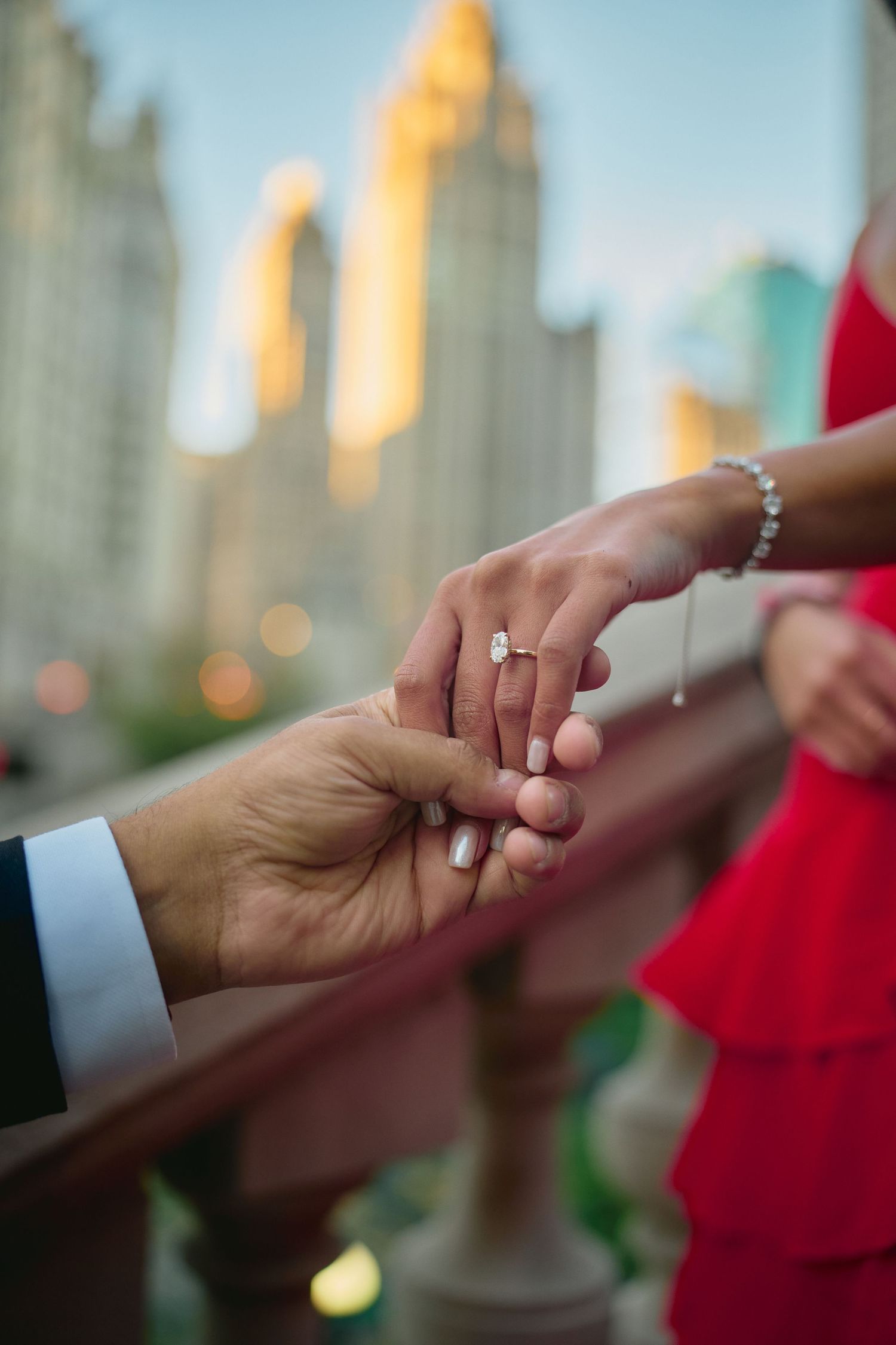 Engagement ring sparkles as hands touch tenderly against a blurred cityscape backdrop at sunset.