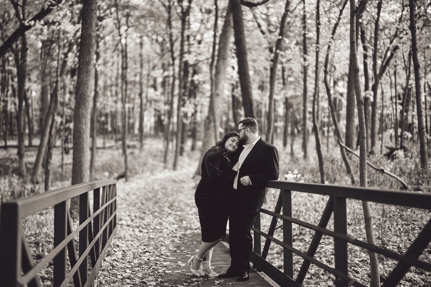 A romantic black and white photo of a couple sharing a tender moment on a wooden bridge in an autumn forest.