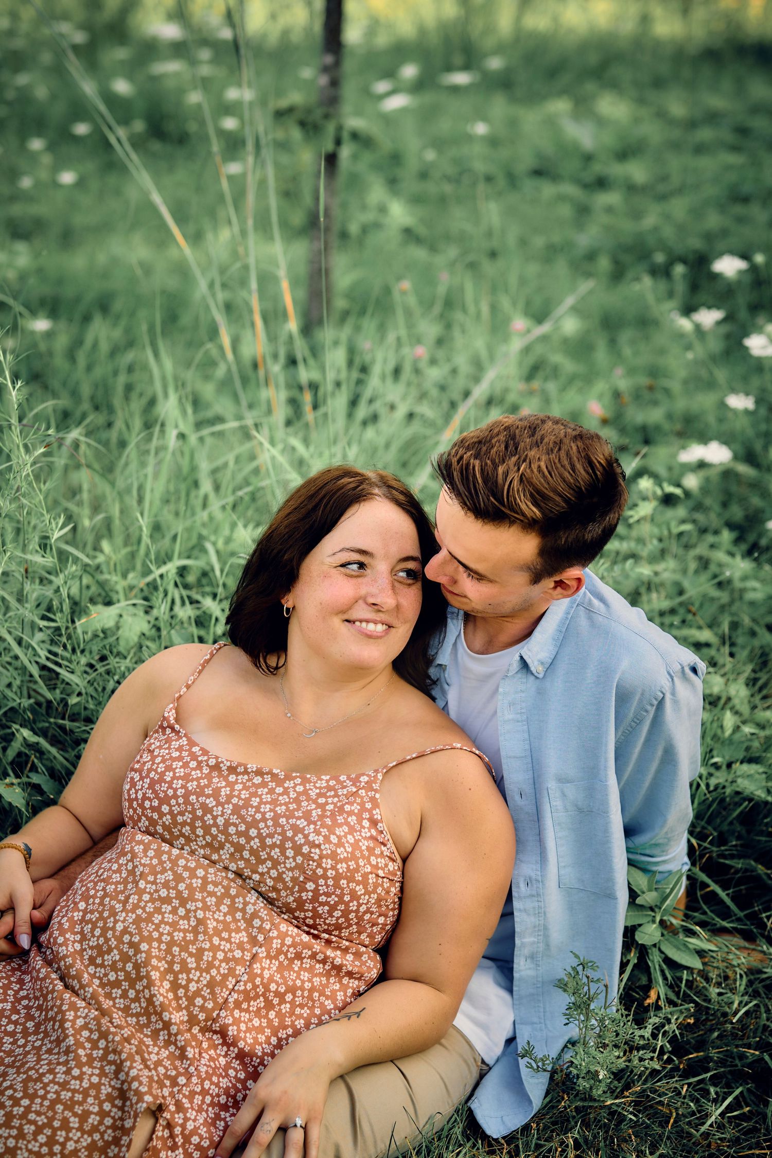 Young couple embraces while sitting in a meadow with wildflowers, sharing an intimate moment on a summer day.
