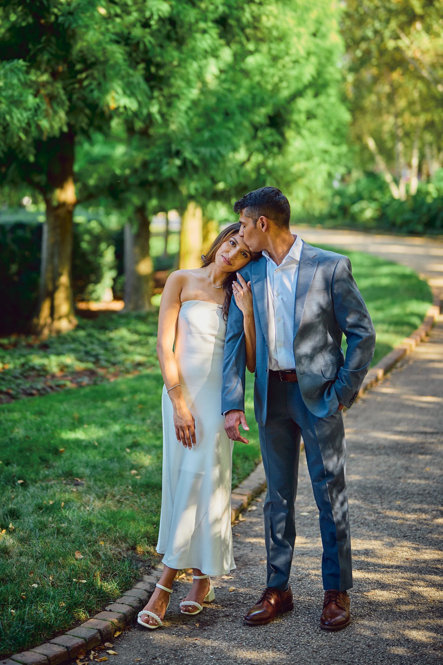 A romantic couple in formal attire share an intimate moment while walking along a tree-lined path on a sunny day.