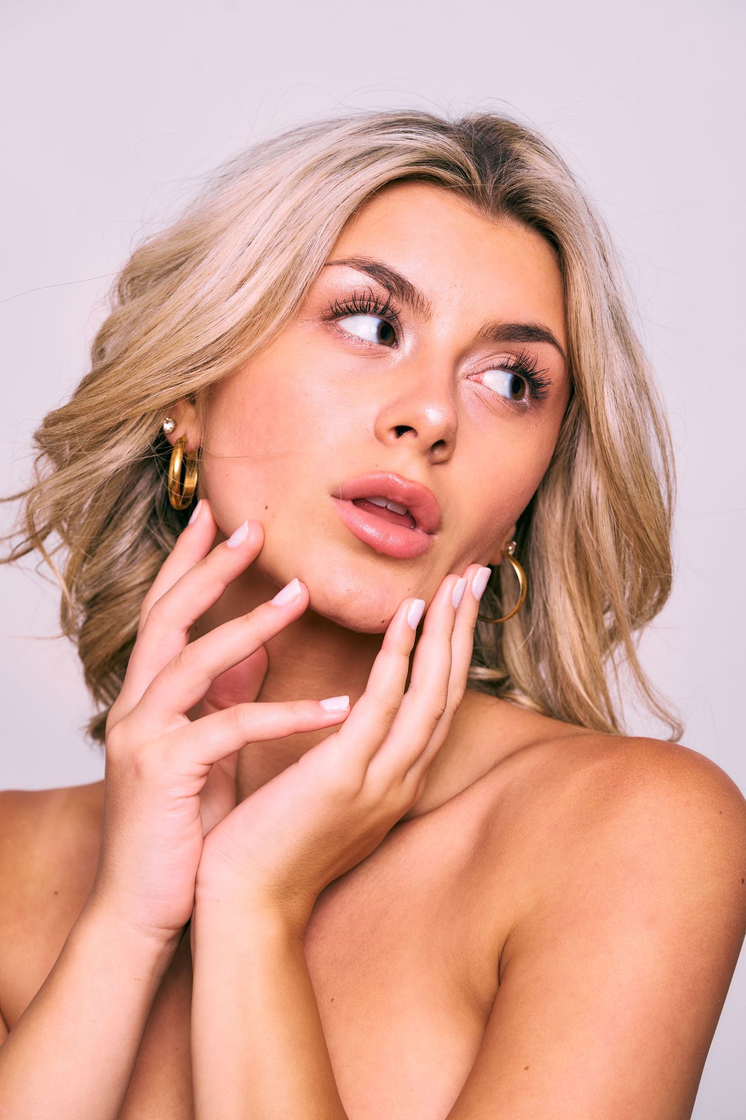 Glamorous beauty portrait with blonde hair, soft makeup, and delicate hand poses against pink background.