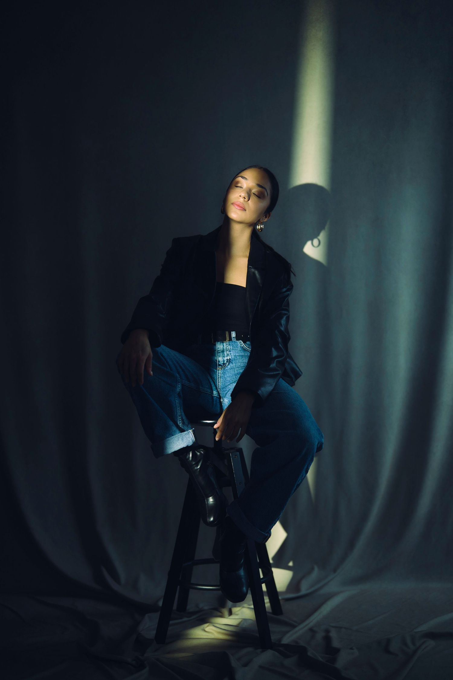 Dramatic fashion portrait with person in black jacket seated on stool under spotlight.
