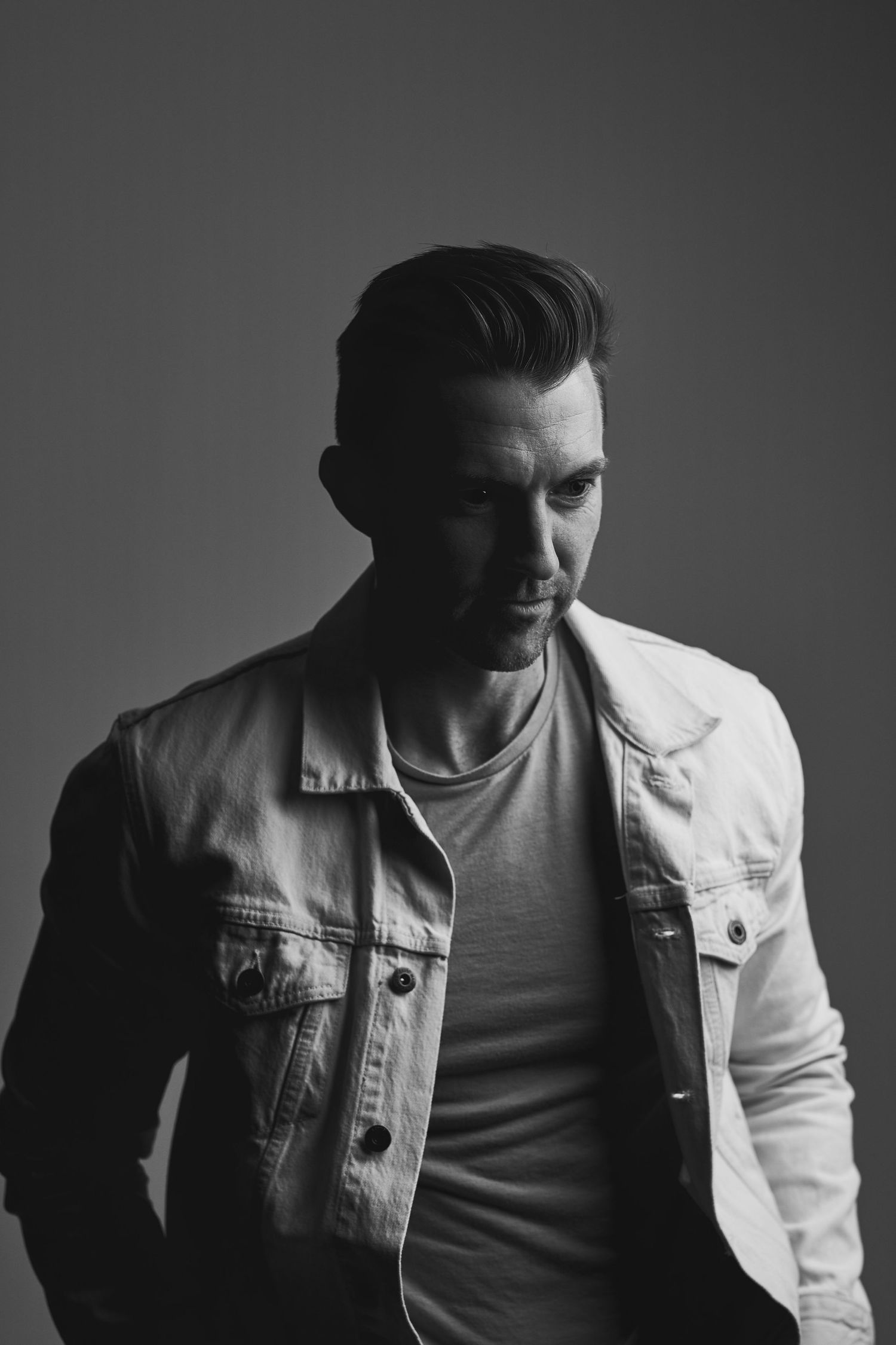 Black and white portrait of man in white denim jacket with styled hair.