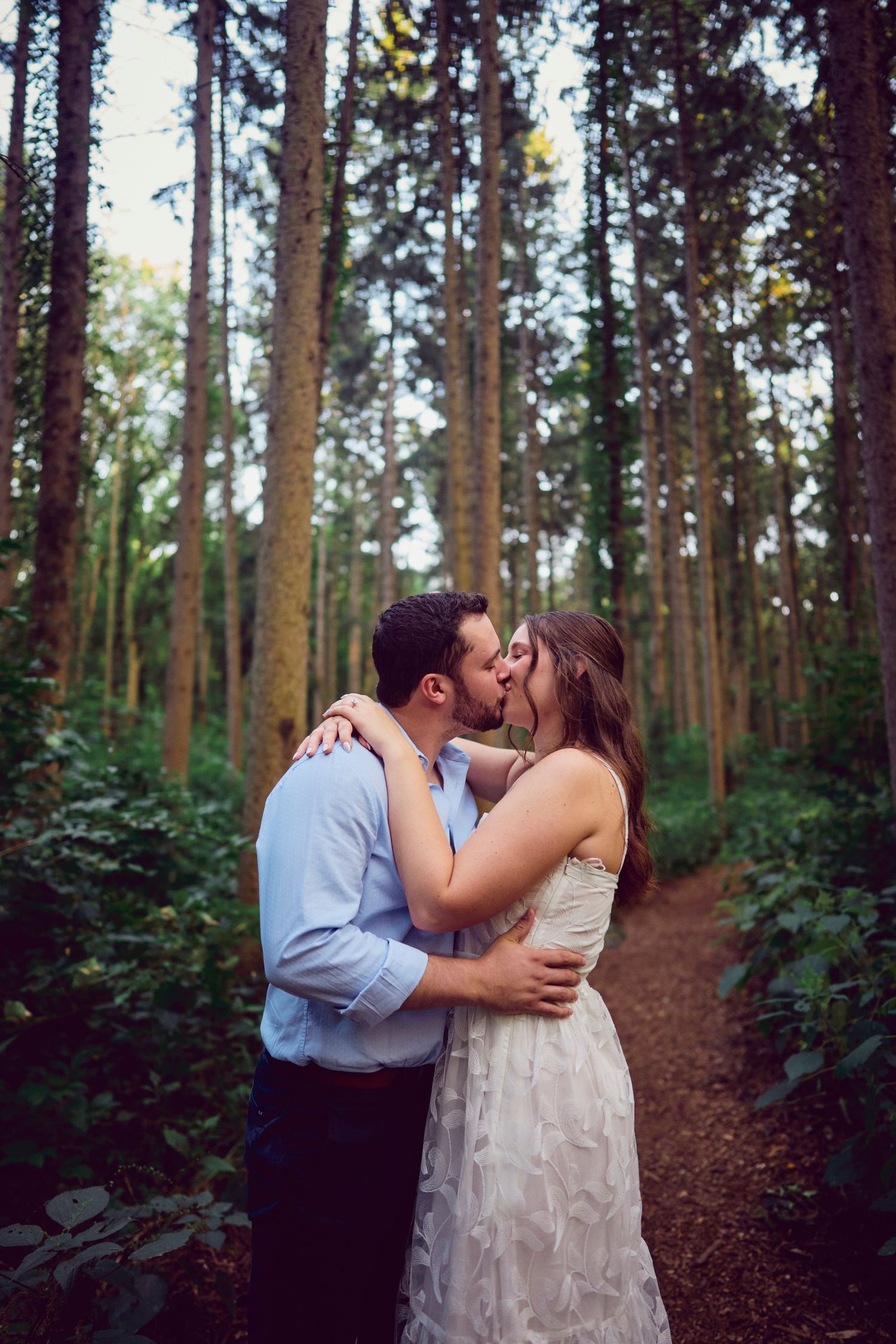 A romantic couple embraces and kisses on a woodland trail surrounded by tall evergreen trees.