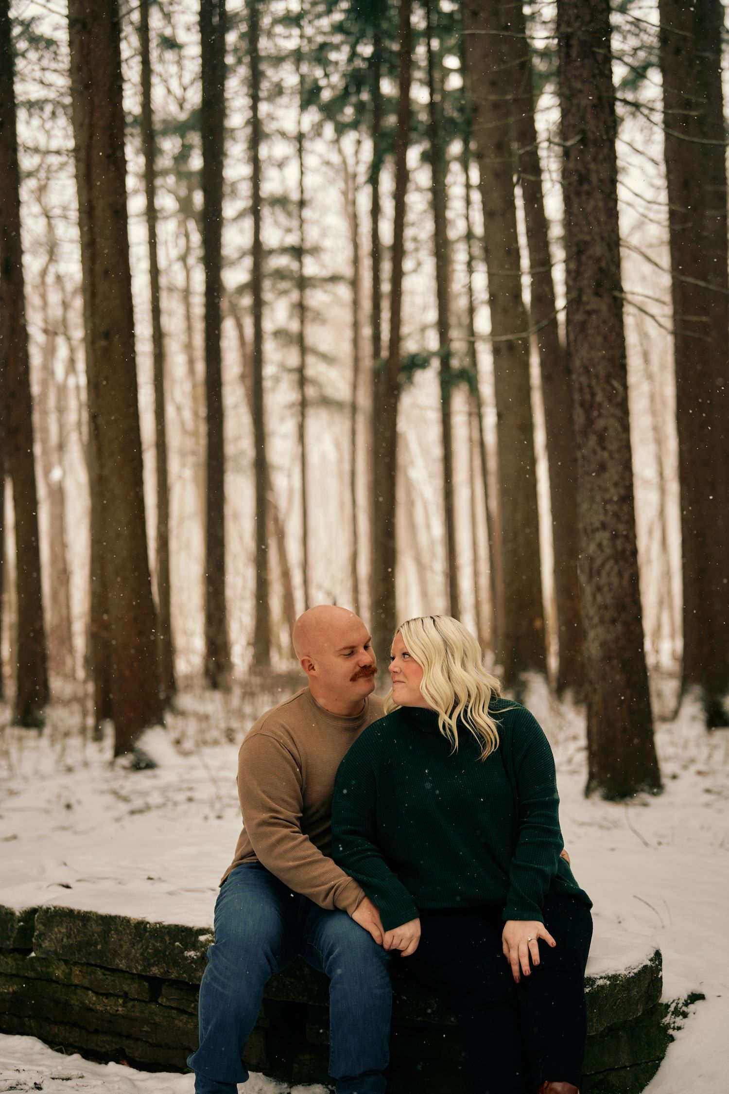 A couple sits together on a fallen log in a snowy winter forest, sharing an intimate moment at sunset.
