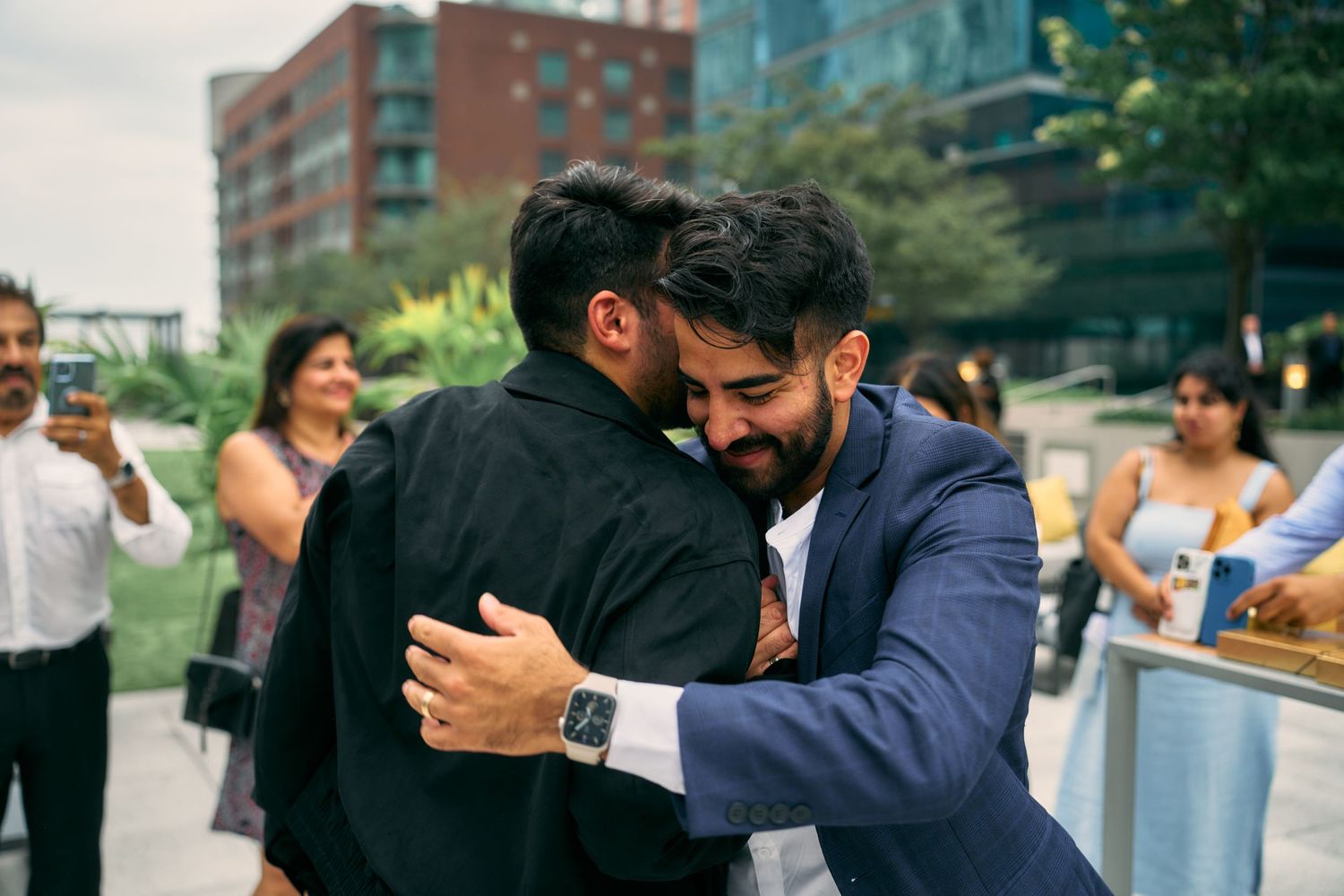 Two men embracing warmly outdoors at a social gathering with an urban building backdrop.