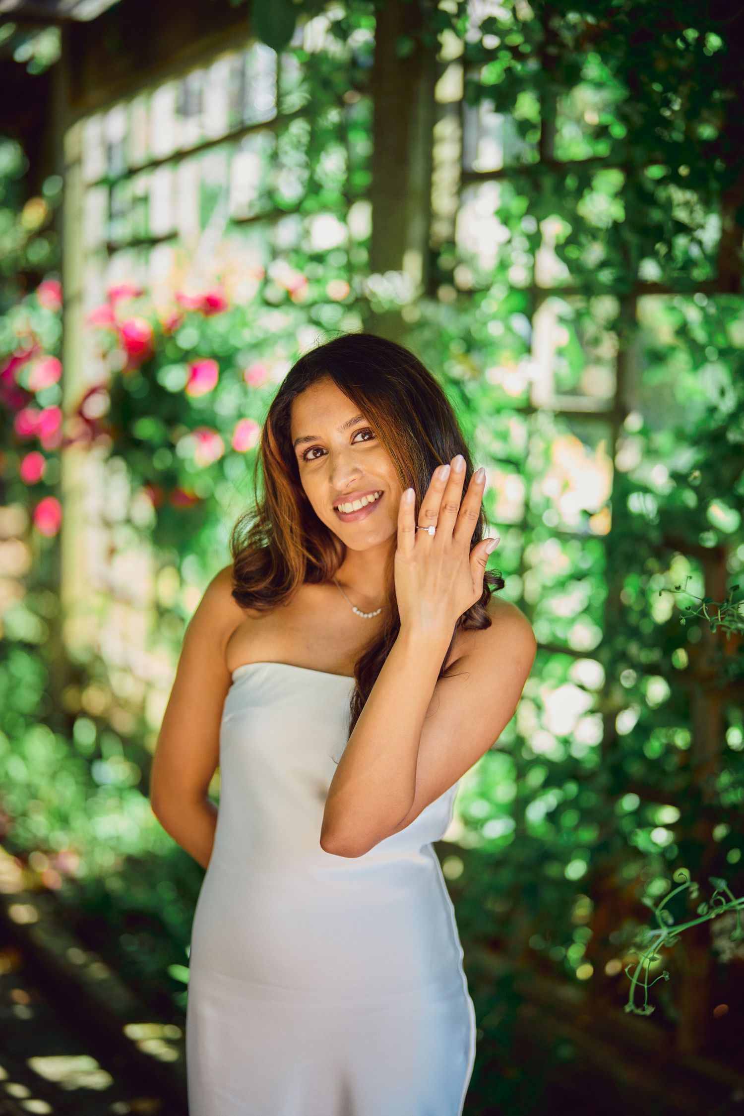 Smiling person in a white strapless dress poses against lush greenery and pink flowers in a garden setting.