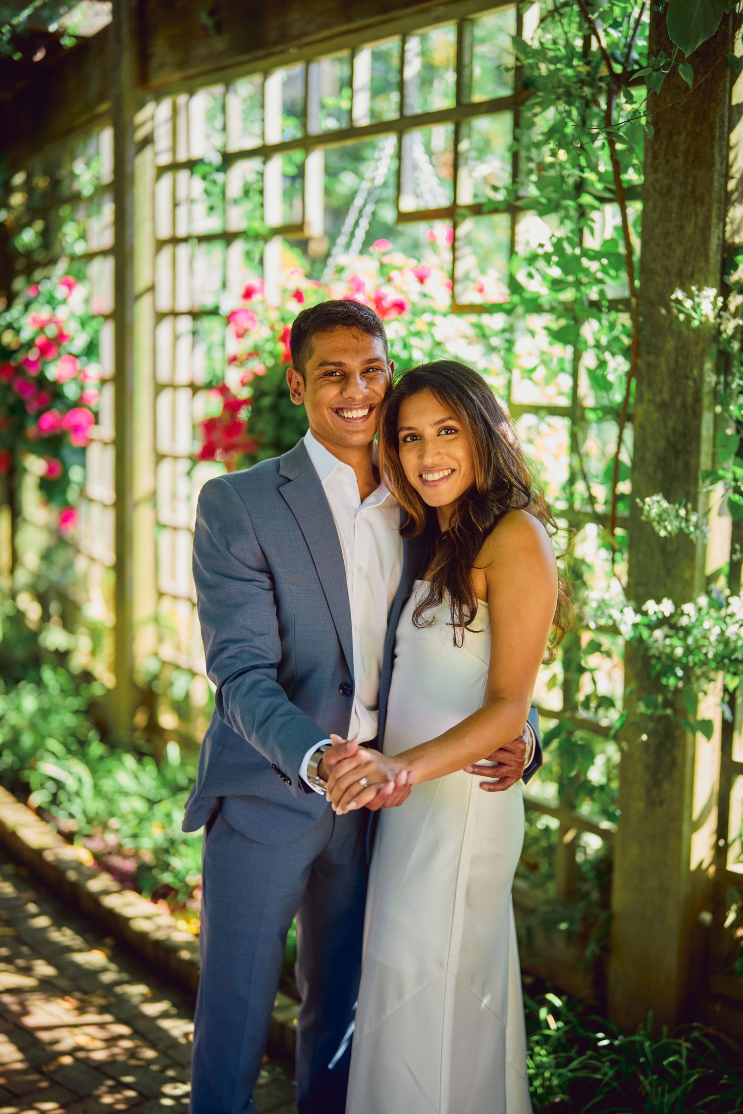 Couple poses for romantic portrait in garden pergola with climbing flowers and dappled sunlight.