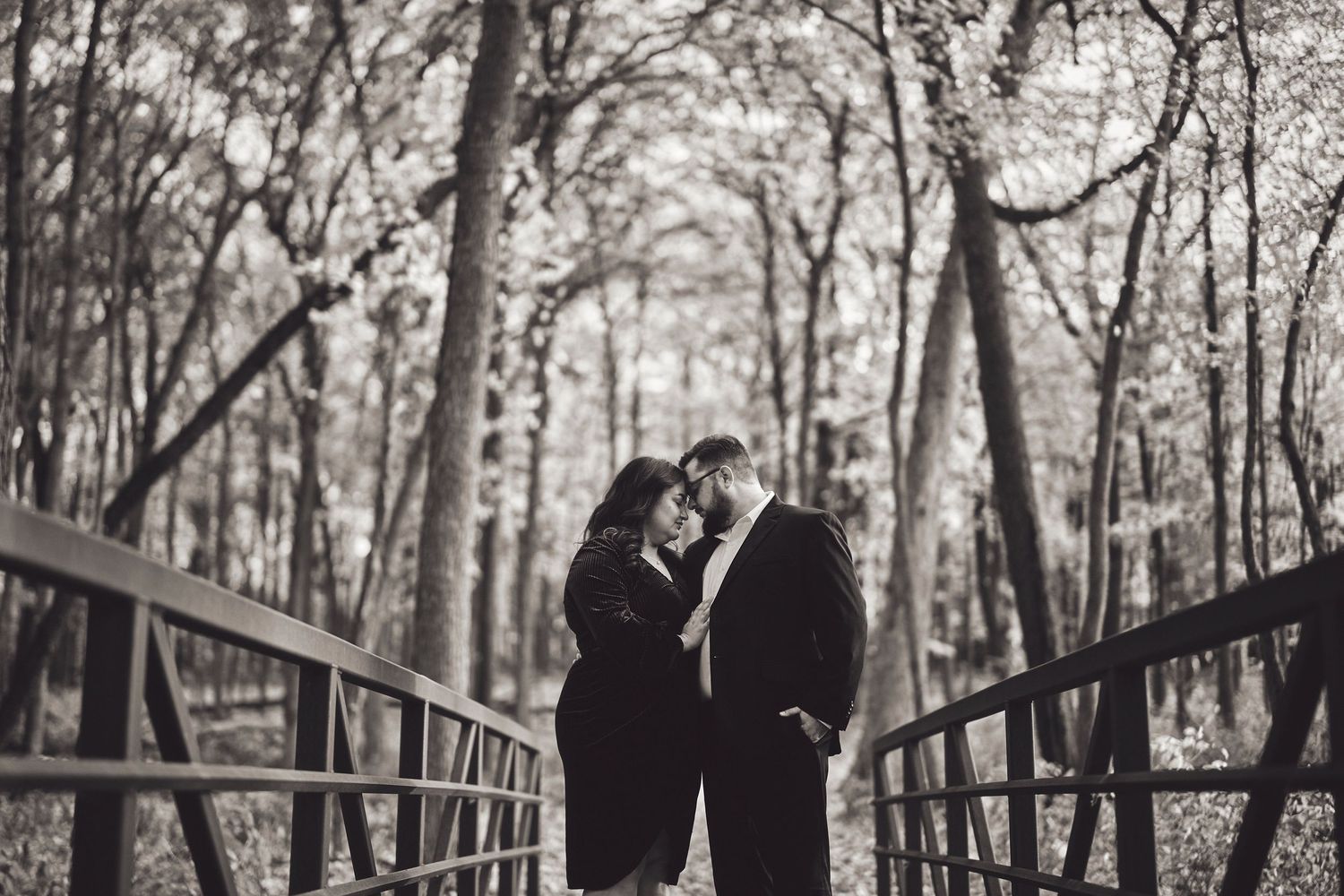 Two people share an intimate moment while standing on a wooden bridge surrounded by a winter forest in black and white.