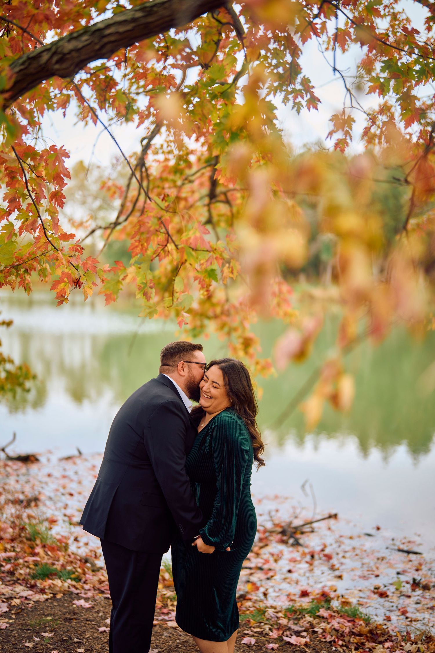 A couple shares a romantic moment by a lake surrounded by autumn foliage with orange and red leaves overhead.