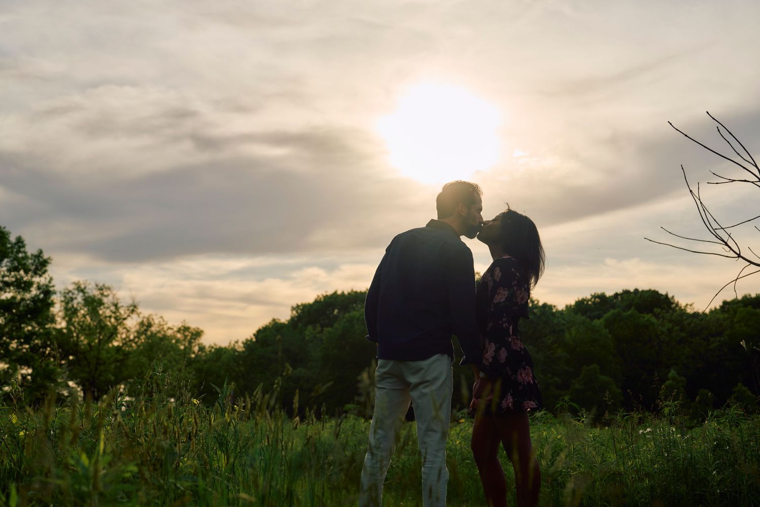 Silhouetted couple stands in a meadow at sunset with trees in the background.
