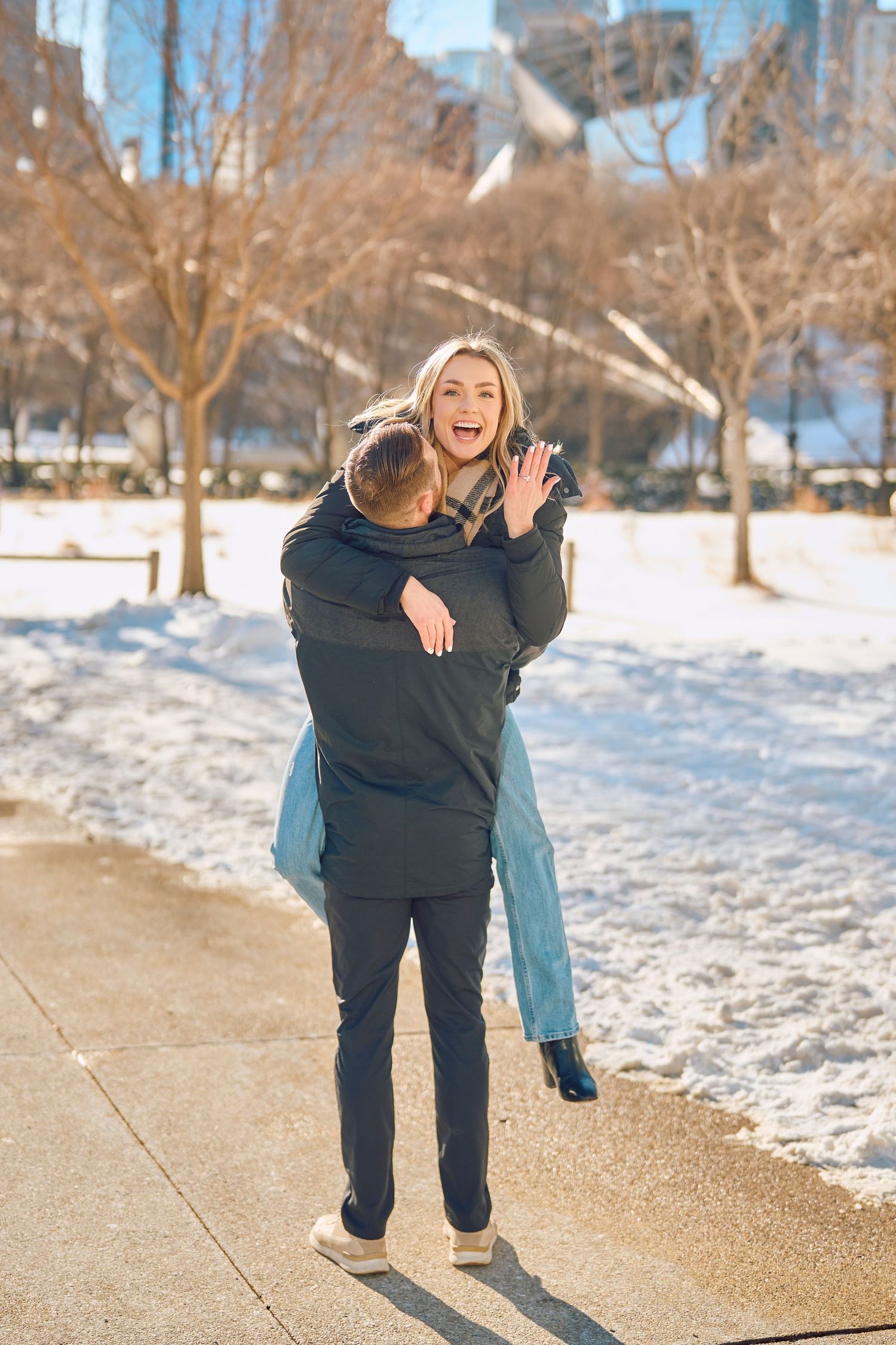 A playful piggyback ride on a snowy winter path with bare trees and urban buildings in the background.