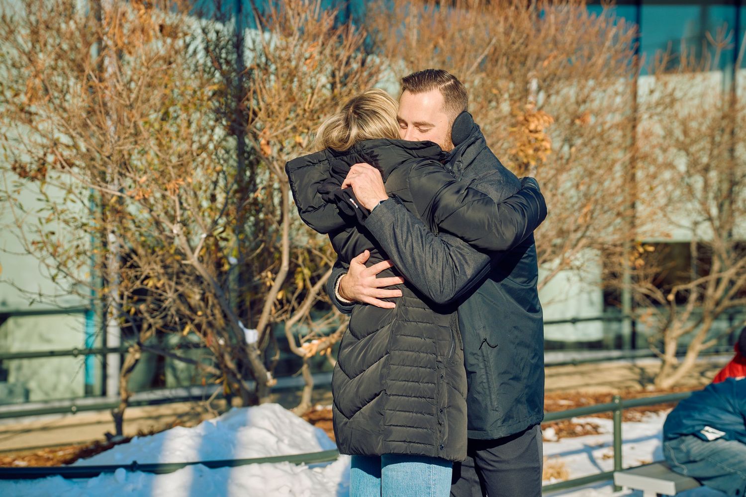Two people embrace in a warm hug on a sunny winter day with bare trees in the background.