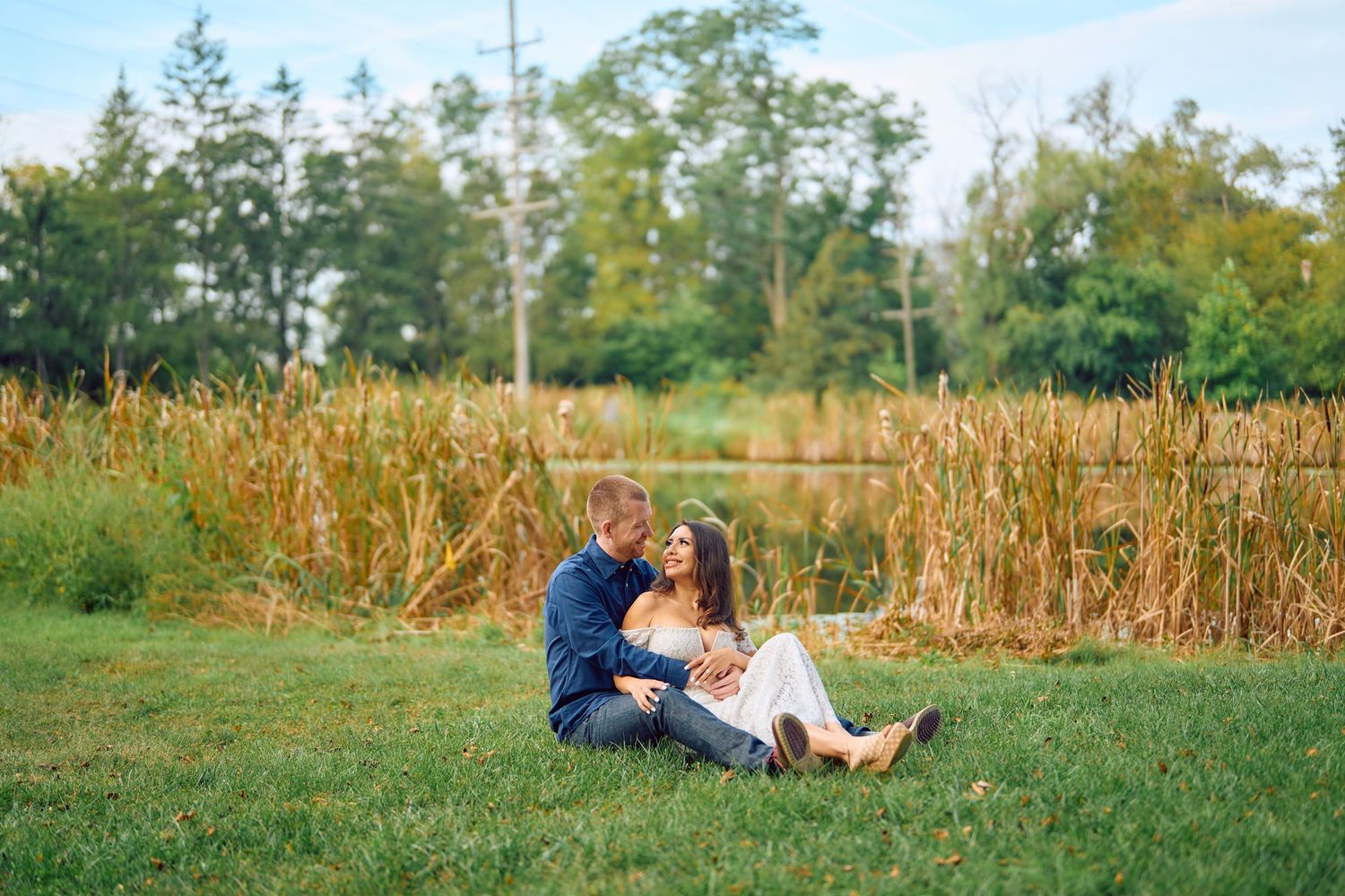 A couple sits together on green grass near tall wheat fields on a sunny summer afternoon.