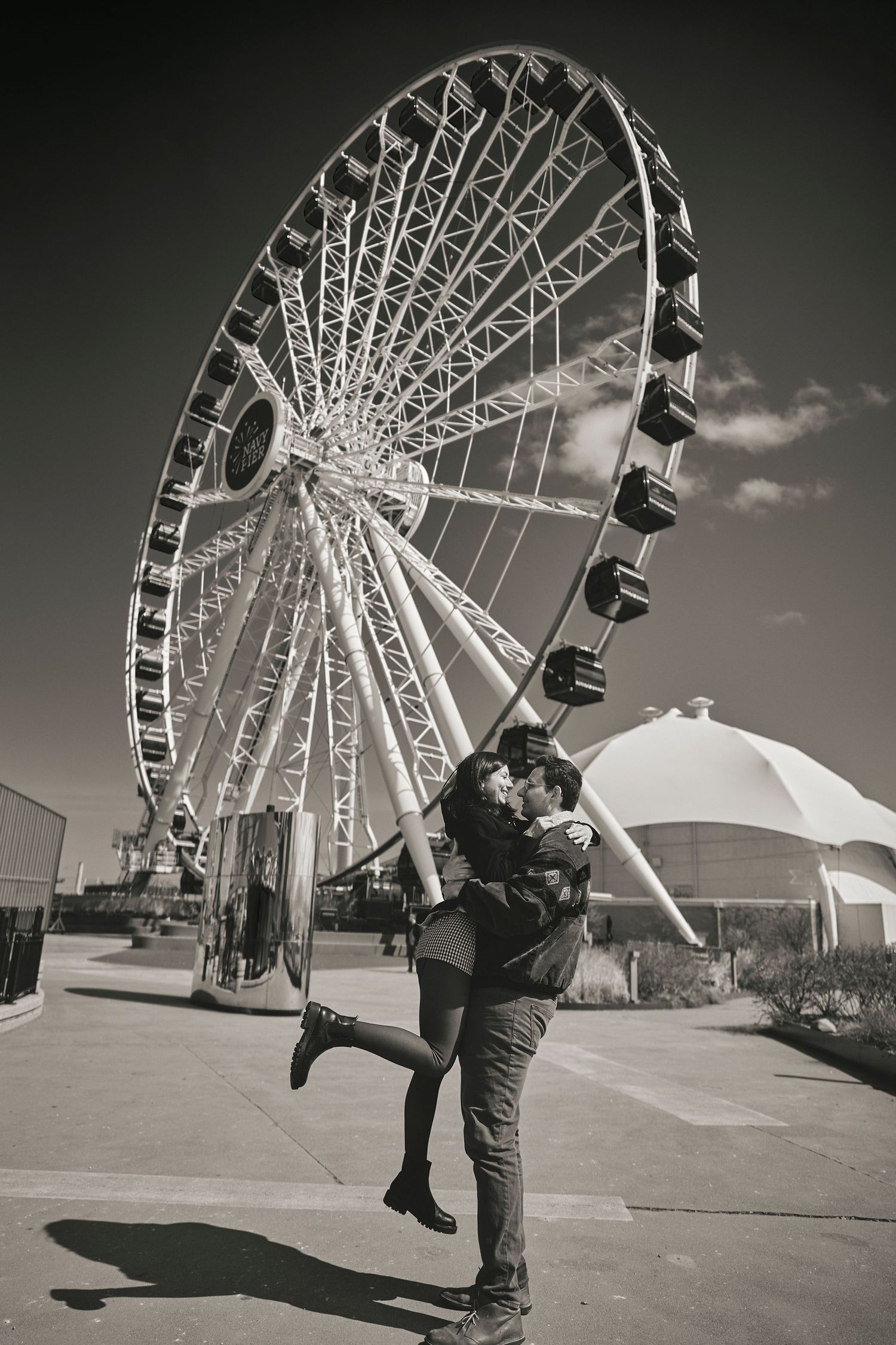 Silhouetted couple embracing romantically in front of a large Ferris wheel under cloudy skies in black and white.