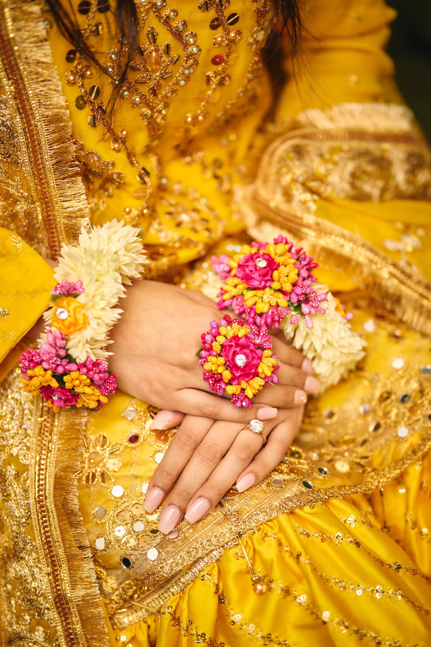 Hands adorned with pink and white floral jewelry rest on a vibrant yellow embroidered traditional Indian wedding dress.