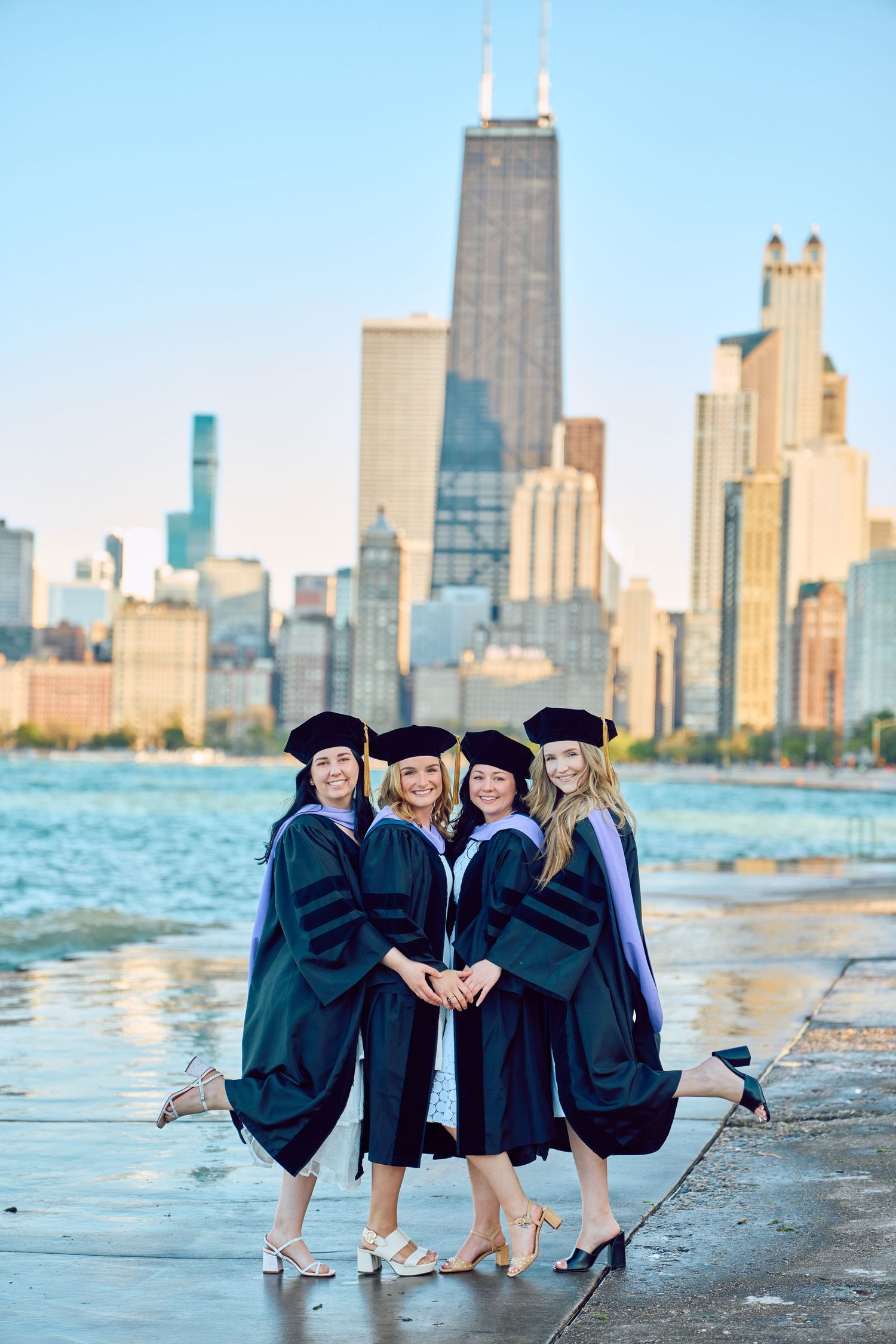 Four graduates in black caps and gowns sit along Lake Michigan with the Chicago skyline in the background.