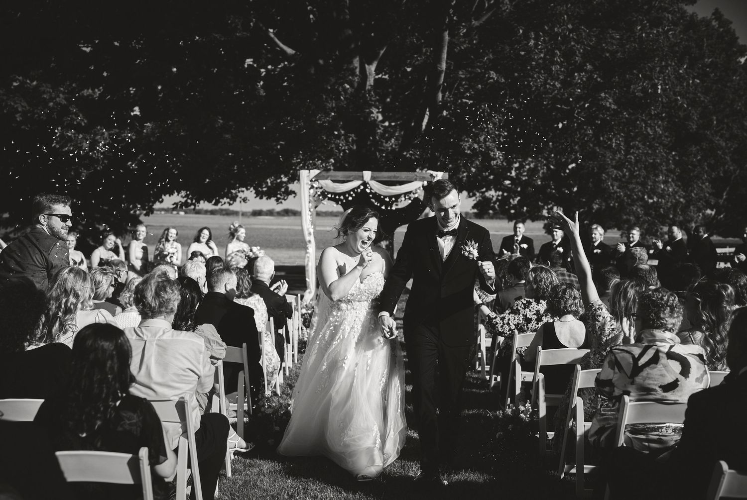Black and white photo of outdoor wedding ceremony with guests under trees.