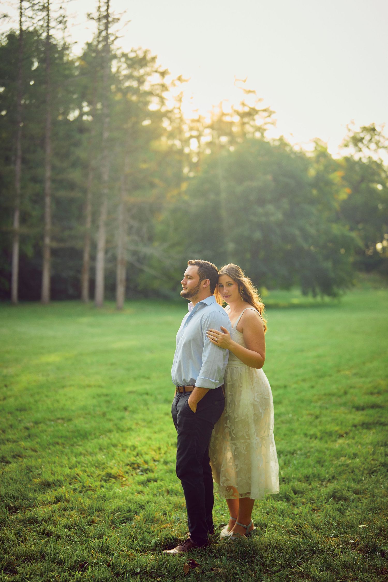 A couple stands together on a grassy lawn at sunset with tall pine trees in the background.