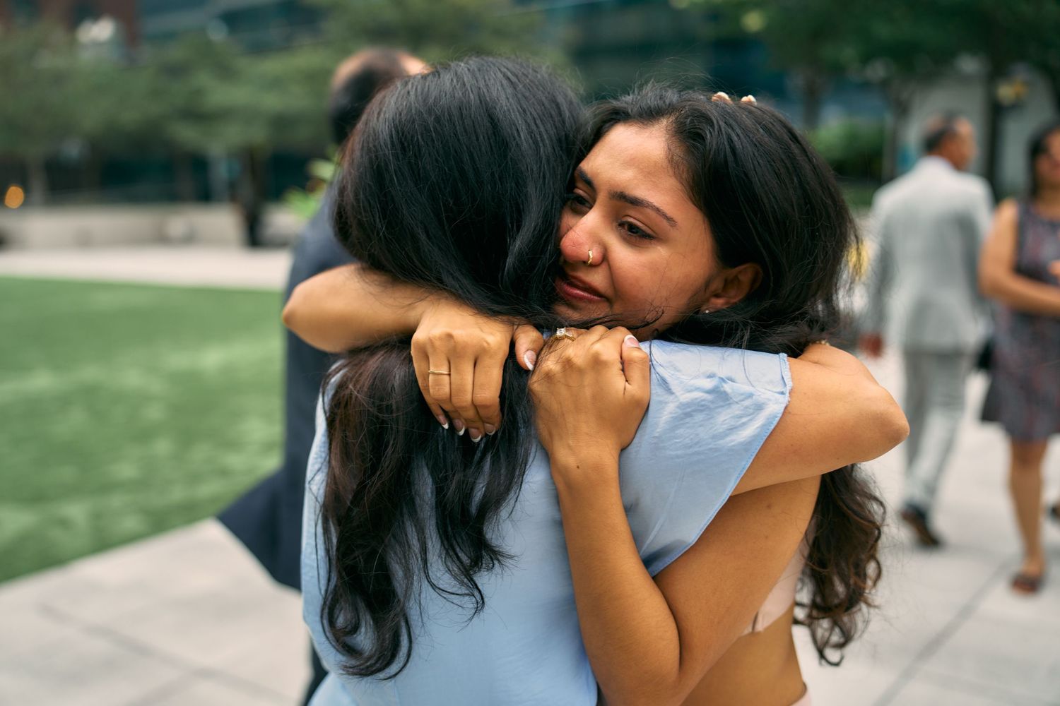 Two friends share an emotional embrace on a sunny day outdoors with green grass in the background.