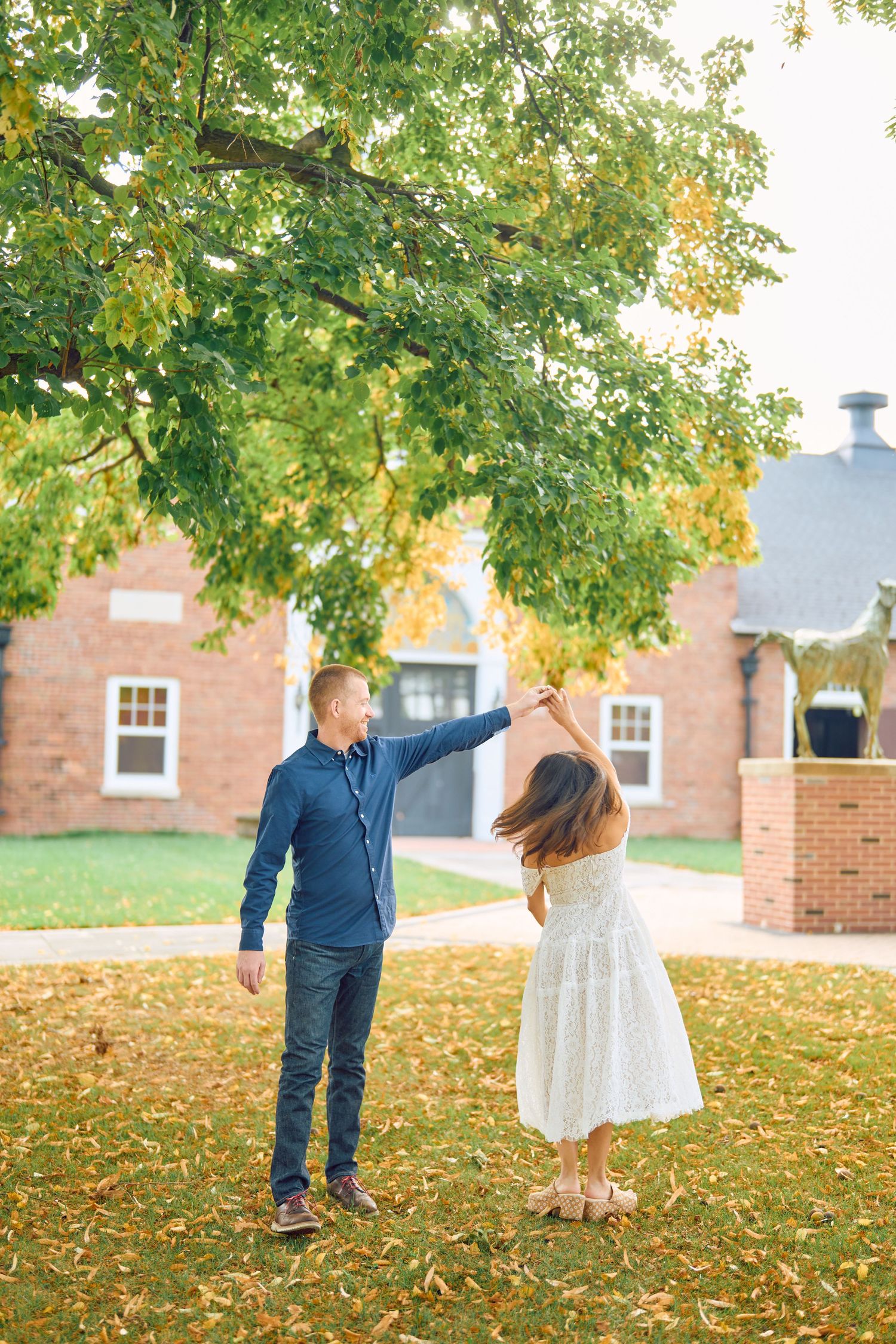 A couple dances together on fallen autumn leaves beneath a large tree outside a brick building.