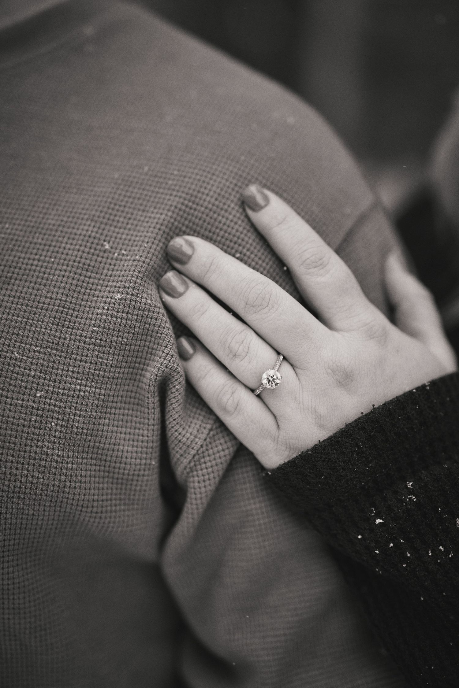 A close-up black and white photo shows a hand wearing an engagement ring resting on textured fabric.