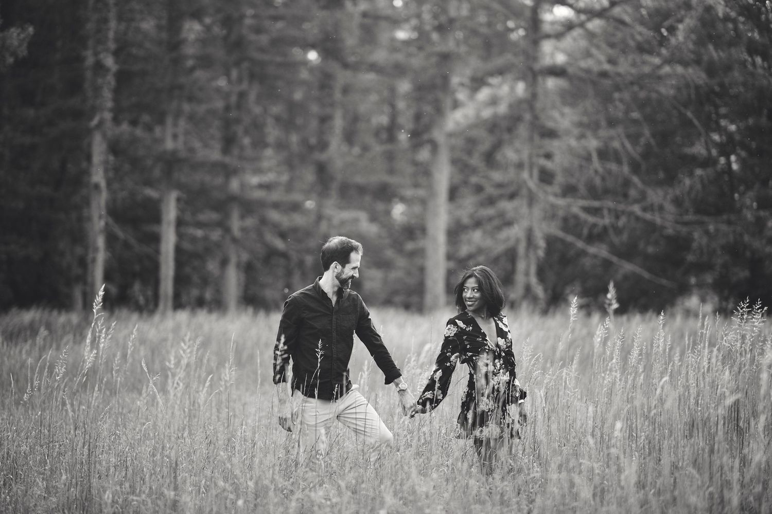 A couple holds hands while walking through tall grass in a forest meadow in black and white.