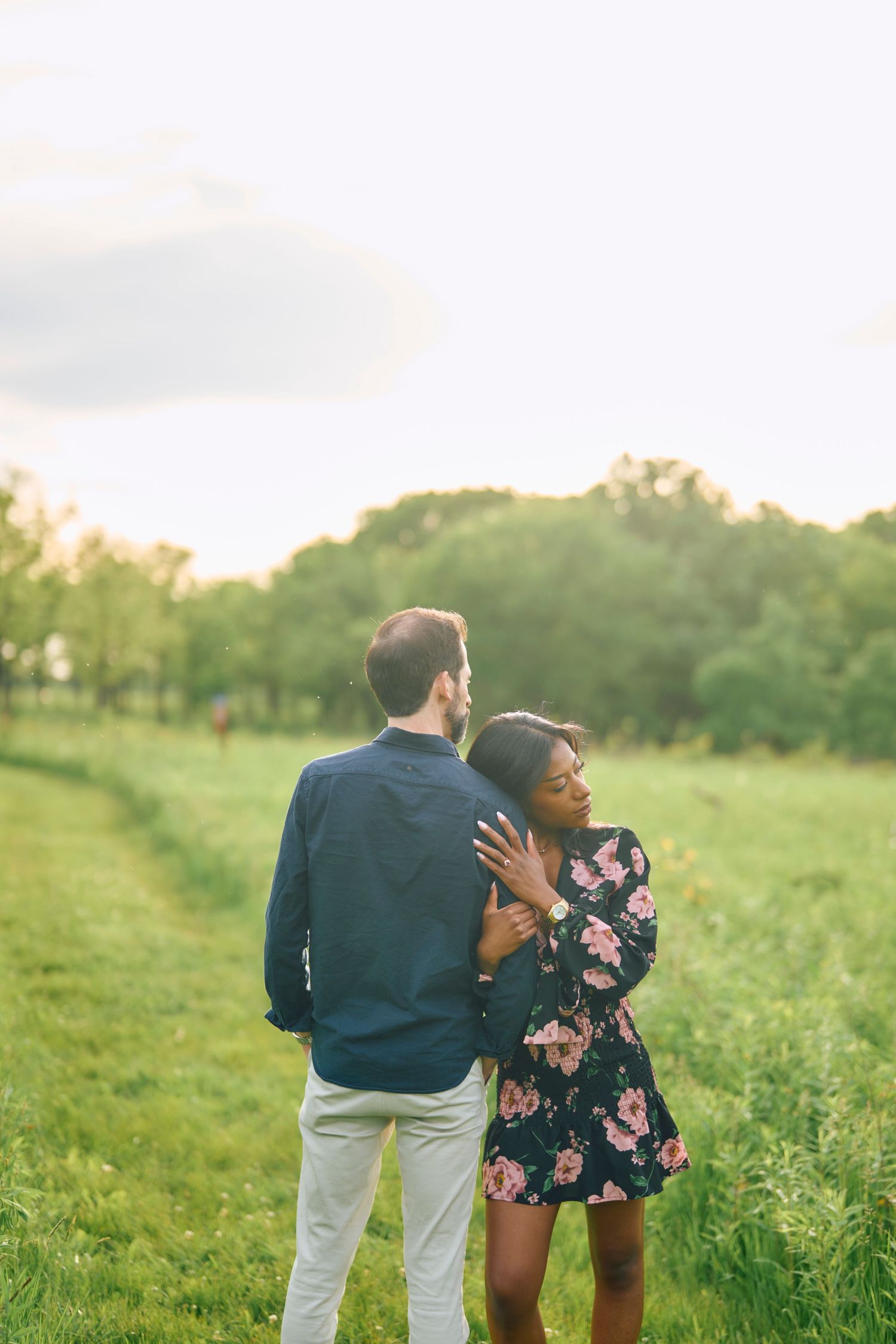 Couple standing together in a romantic pose on a grassy hill during golden hour with trees in background.