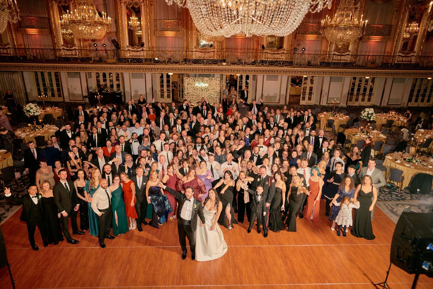 Large group photo of wedding guests gathered in an ornate ballroom with crystal chandeliers and elegant decor.