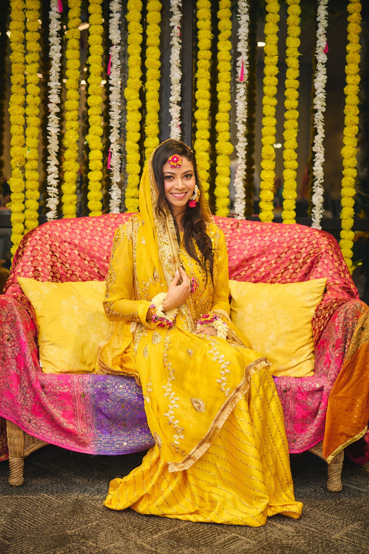 A person wearing a bright yellow traditional Indian outfit poses against a backdrop of marigold flower decorations.