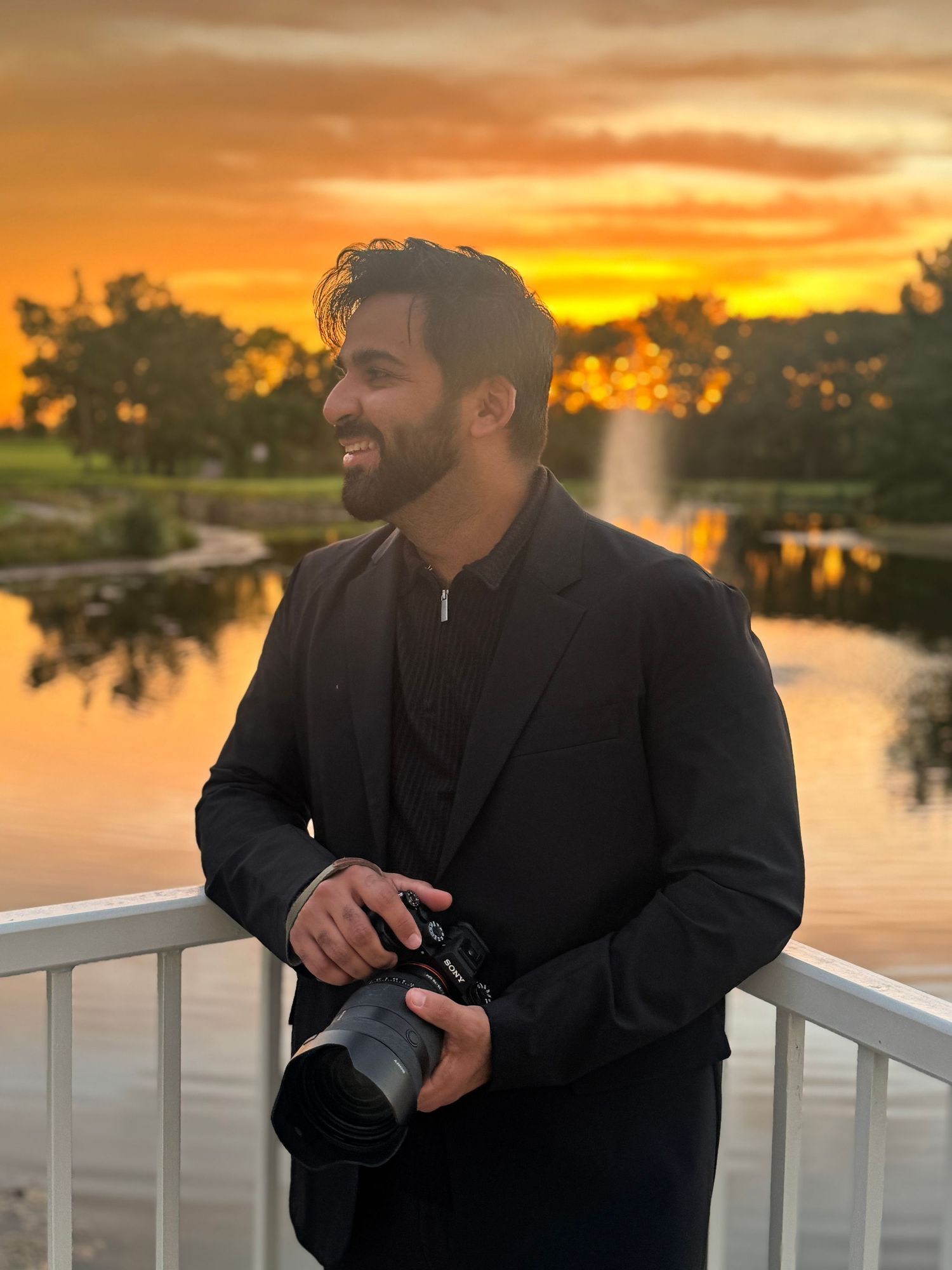 Man in black blazer holding camera smiles at golden sunset over pond with fountain and trees.