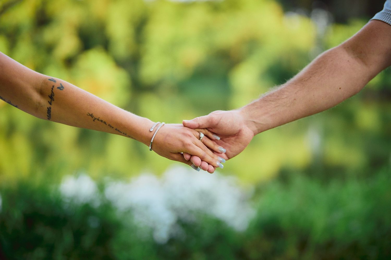 Two hands with tattoos lovingly hold each other in a tender moment against a soft green natural background.