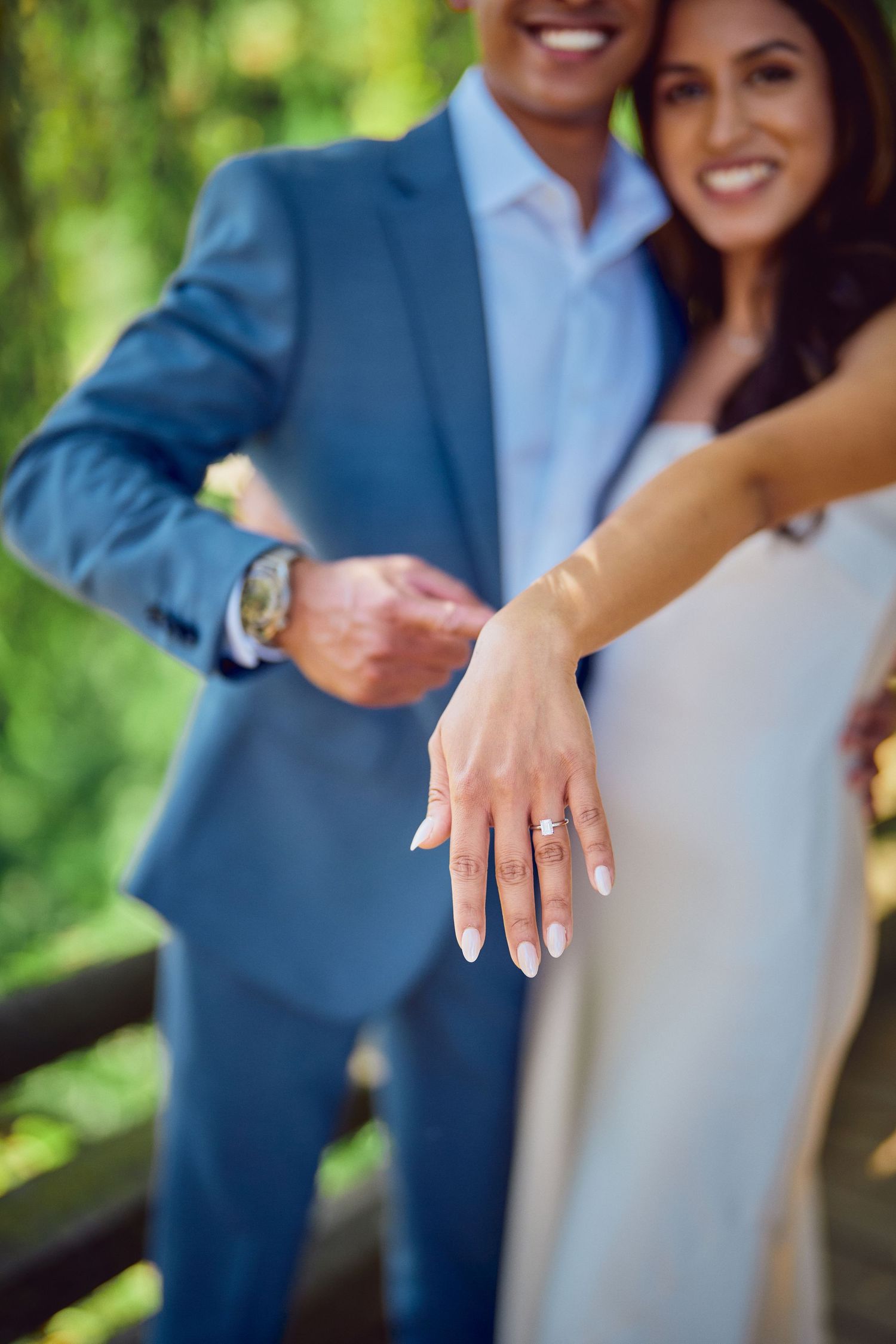 A close-up photograph of an engagement ring on a hand, with a blue suit and light dress visible in the background.