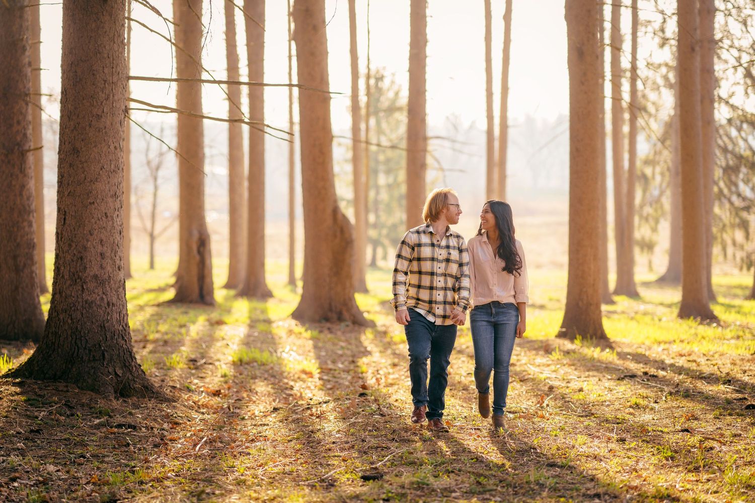 A couple walks together hand in hand through a sunlit forest during golden hour as rays shine between tall trees.