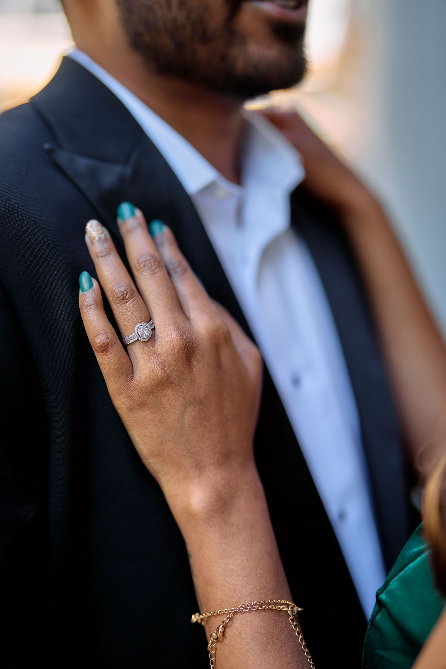 A hand wearing a delicate diamond engagement ring touches a suit lapel, with a gold bracelet adorning the wrist.
