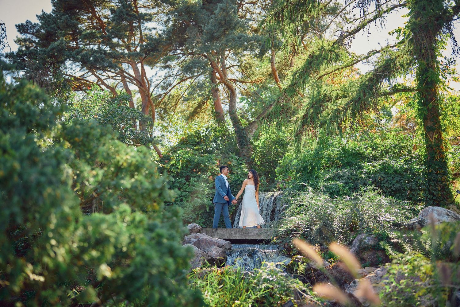 A couple stands together on a stone pathway surrounded by lush evergreen trees and natural foliage in a garden setting.